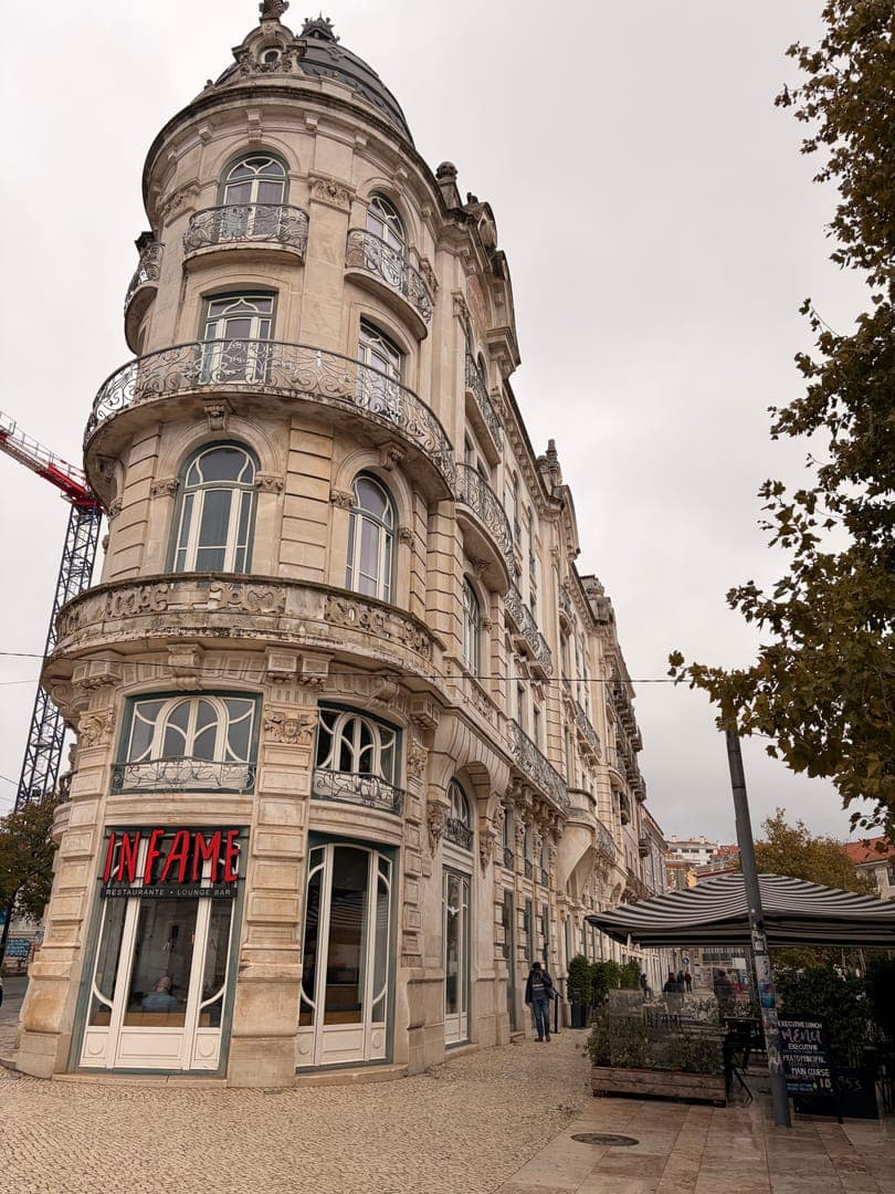 This captivating low-angle shot showcases a grand, ornate building with a rounded corner and intricate balconies, exuding a timeless European charm. The ground floor features a restaurant named 'INFAME RESTAURANTE LOUNGE BAR' with a striking red neon sign, inviting passersby into its elegant interior. The scene is completed by a quaint outdoor seating area with striped awnings and lush greenery, hinting at a relaxed urban atmosphere under a soft, overcast sky.