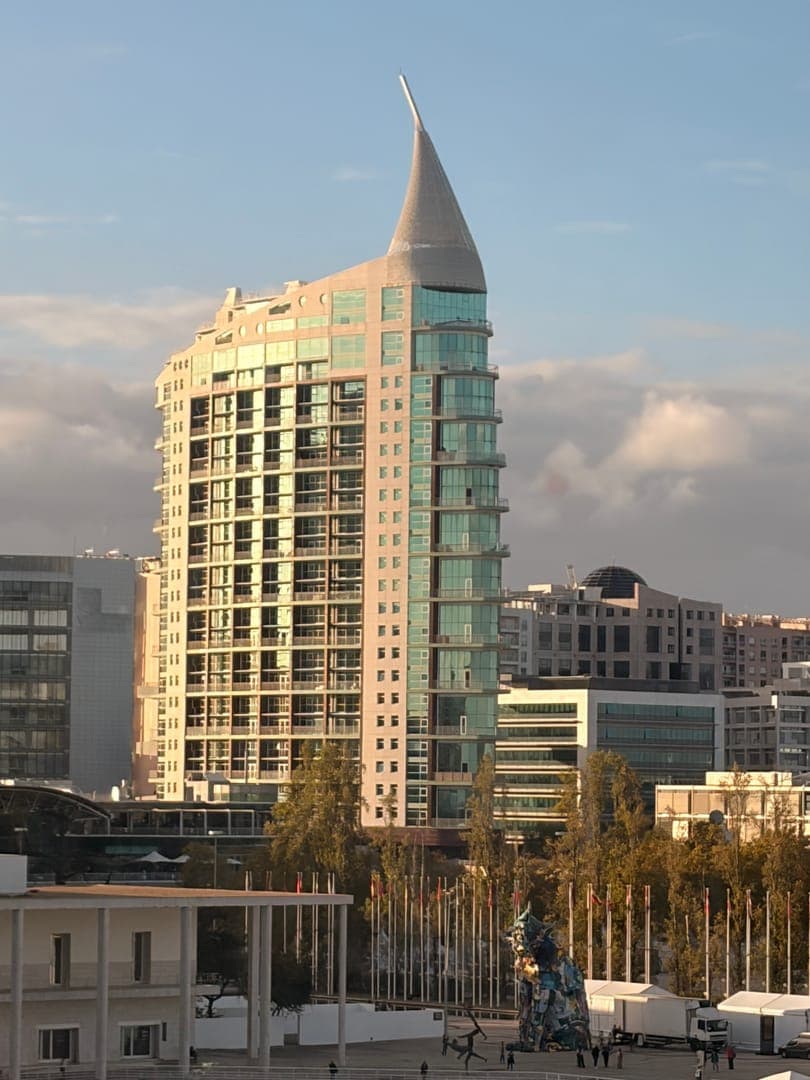 This striking image captures the iconic São Gabriel Tower in Lisbon, Portugal, with its distinctive conical spire reaching towards a partly cloudy sky. The modern architecture, featuring a mix of glass and light-colored facades, stands tall amidst other urban buildings and a tree-lined plaza below. The warm, golden hour light bathes the scene, highlighting the building's unique design and creating a serene yet vibrant city atmosphere.