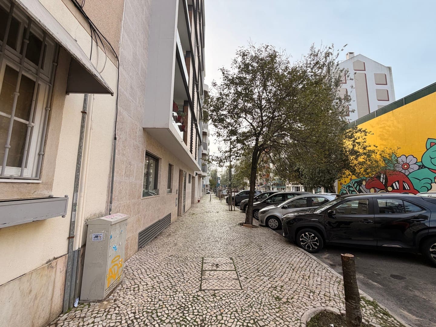 A charming cobblestone sidewalk winds alongside a row of parked cars and buildings, leading the eye towards a vibrant yellow wall adorned with playful street art. The scene captures a quiet urban moment, with trees lining the path and a mix of modern and traditional architecture creating an interesting contrast. The overall mood is one of everyday city life, with a touch of artistic flair adding character to the street.