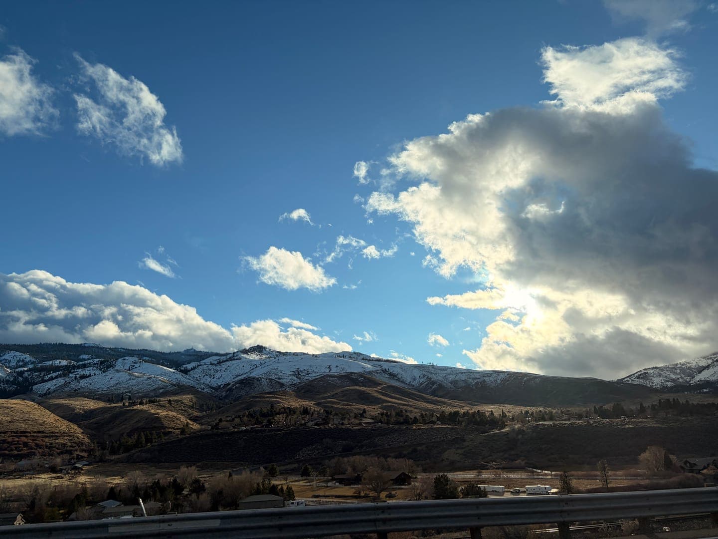 A breathtaking view of snow-capped mountains under a dramatic sky, where patches of bright blue peek through fluffy white and dark, brooding clouds. The sun casts a warm glow on the golden-brown foothills, creating a striking contrast with the cool, snowy peaks. This scenic landscape captures the serene beauty of nature's ever-changing canvas.
