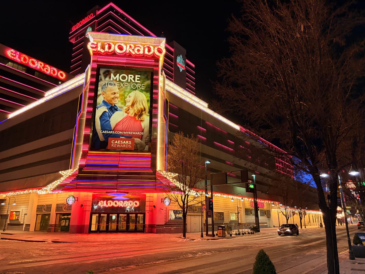 The Eldorado Casino lights up the night with its vibrant red neon signs, prominently displaying 'ELDORADO' on its facade and a large billboard advertising 'MORE TO EXPLORE' with a happy couple. The street below is dusted with snow, reflecting the glow of the casino and streetlights, creating a festive yet serene winter evening atmosphere. A few cars drive by, and bare trees line the sidewalk, adding to the cool, crisp feel of the scene.