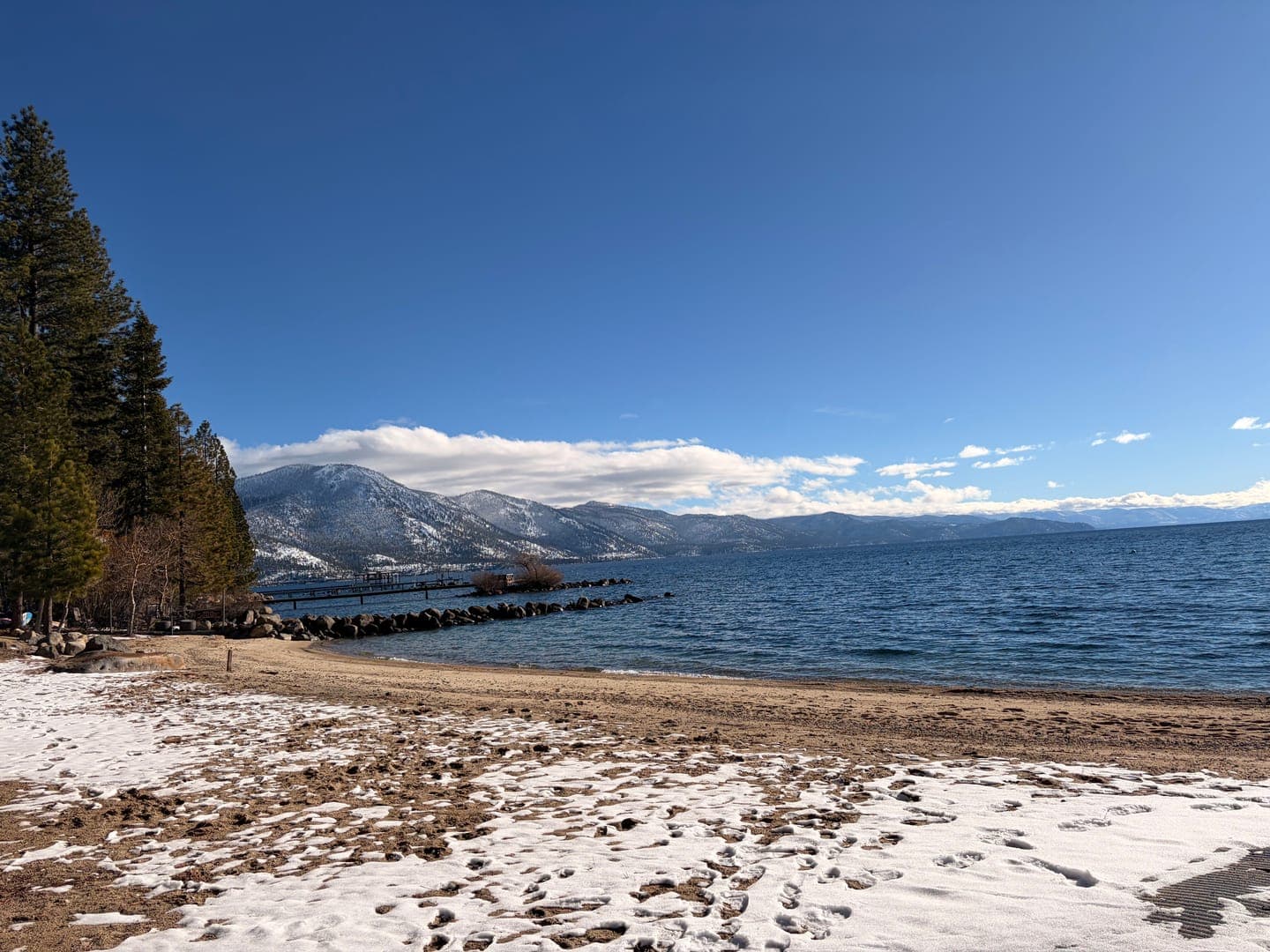 A serene winter scene unfolds at Lake Tahoe, where patches of snow dot the sandy beach in the foreground. Towering pine trees line the left side, leading the eye towards the vast expanse of the deep blue lake. In the distance, majestic snow-capped mountains rise under a clear, bright blue sky, creating a breathtaking backdrop.