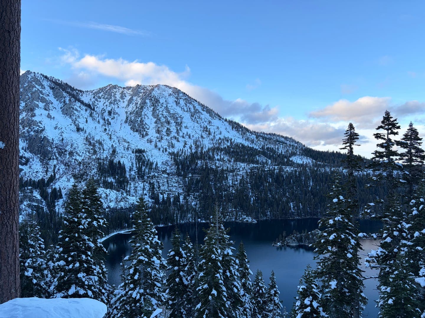 A breathtaking winter wonderland unfolds with snow-capped mountains towering over a serene, dark blue lake. Pine trees, dusted with fresh snow, frame the foreground, adding to the tranquil and majestic mood of the scene. The clear blue sky with scattered clouds completes this picturesque view, inviting viewers to immerse themselves in nature's beauty.