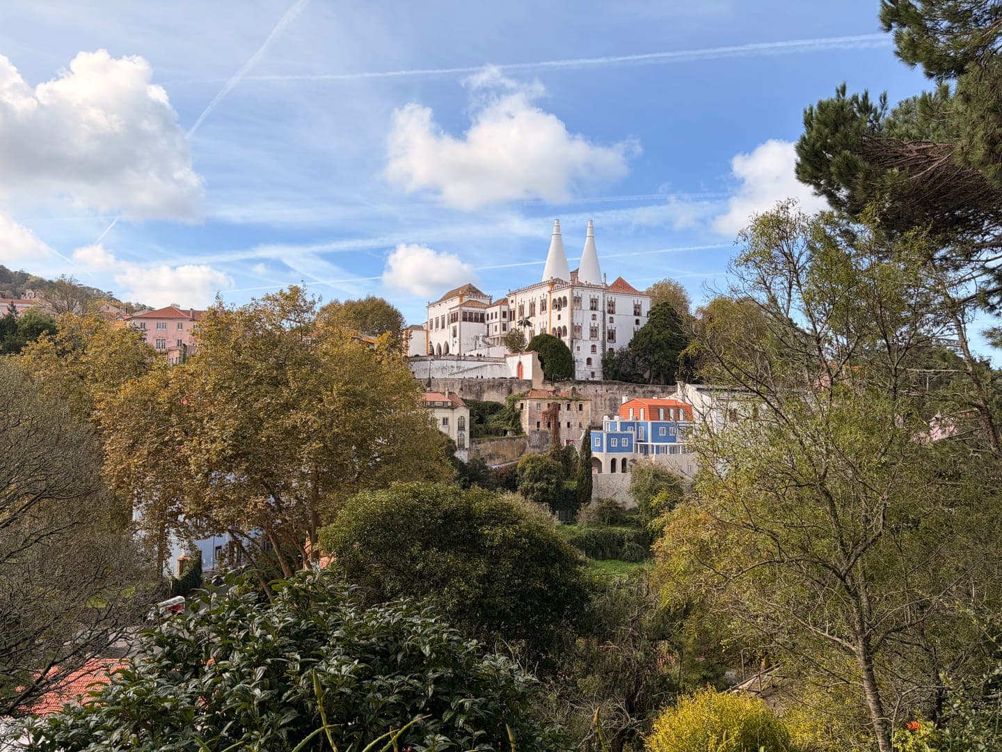 A picturesque view of the National Palace of Sintra, Portugal, with its iconic twin conical chimneys, rises above a lush green landscape. The charming town buildings with their colorful roofs are nestled among the trees, creating a vibrant and historic scene under a bright blue sky with wispy clouds and contrails. This enchanting vista captures the timeless beauty and architectural grandeur of Sintra.