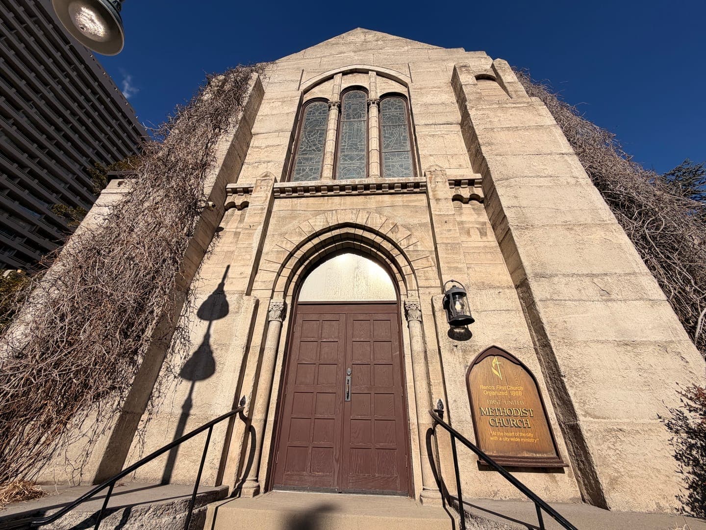This low-angle shot captures the imposing facade of a historic Methodist Church, its stone walls partially covered in dry, climbing vines. The warm sunlight casts dramatic shadows from an overhead light fixture and a decorative lantern onto the textured stone, creating a sense of depth and age. A wooden sign near the entrance clearly identifies it as a 'METHODIST CHURCH,' adding to the building's timeless and reverent atmosphere.