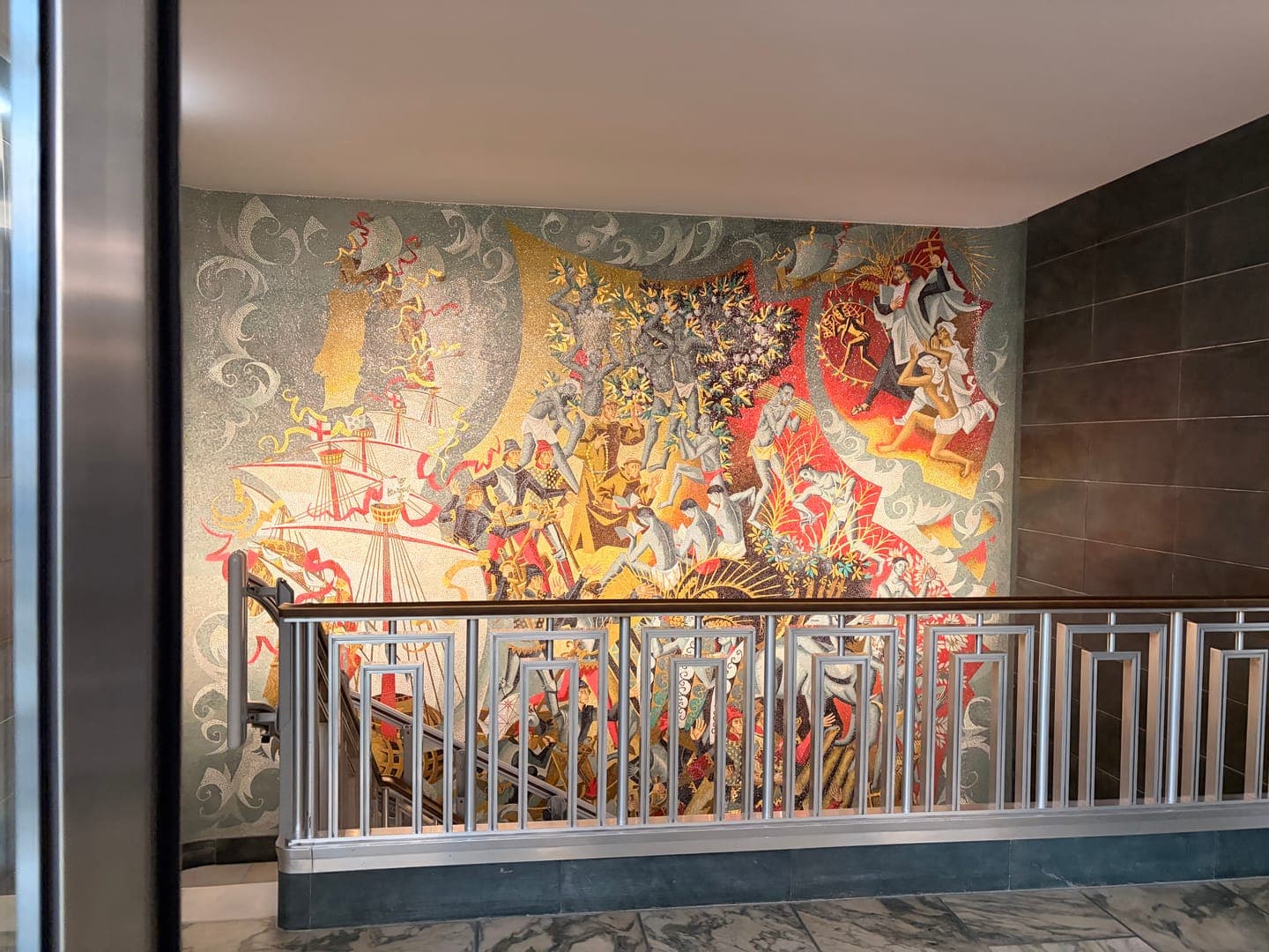 This vibrant mosaic mural, rich in detail and color, depicts a dynamic historical scene with ships, figures in period attire, and dramatic action. The artwork, possibly illustrating a significant event or narrative, is framed by a stylish art deco-inspired railing, adding to the grandeur of the space. The intricate tile work and the energetic composition create a captivating visual experience, inviting viewers to explore its many layers.