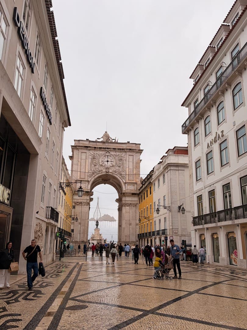 Strolling through the historic streets of Lisbon, Portugal, we're greeted by the majestic Rua Augusta Arch, a grand gateway to the city's vibrant heart. The intricate mosaic pavement leads us toward the arch, with charming buildings like 'DESIGN' and 'ANDAZ' lining the bustling street. The atmosphere is lively yet relaxed, as people wander and enjoy the architectural beauty and unique urban landscape.