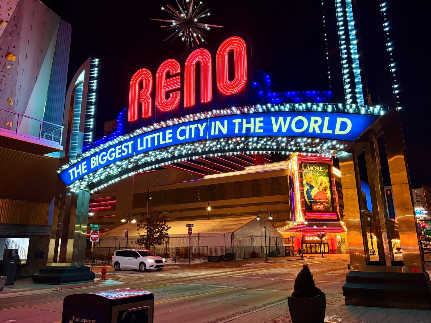 The iconic Reno Arch, brightly lit with neon red and blue, proudly proclaims 'THE BIGGEST LITTLE CITY IN THE WORLD' against the night sky. The vibrant lights reflect off the wet street, creating a dazzling and inviting atmosphere. In the background, other illuminated buildings and a white car add to the lively urban scene of downtown Reno.