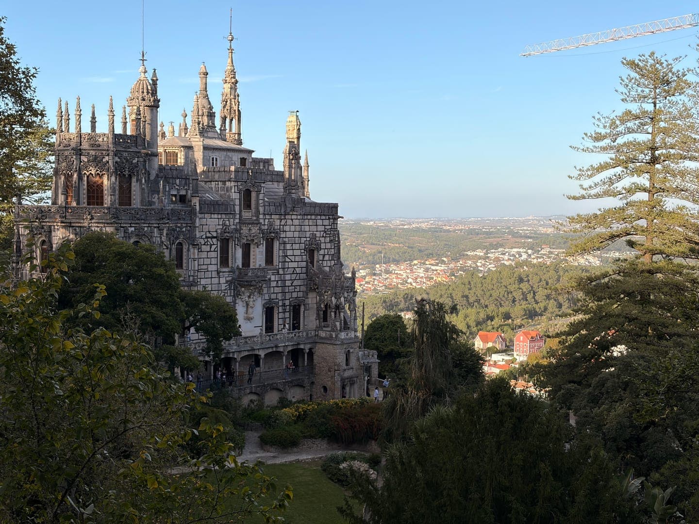 Perched majestically amidst lush greenery, the Quinta da Regaleira palace in Sintra, Portugal, showcases its intricate Gothic architecture under a clear sky. The ornate details of the palace, with its numerous spires and decorative stonework, stand out against the sprawling landscape of the town below. This enchanting view captures the historical grandeur and picturesque surroundings of one of Portugal's most captivating estates.