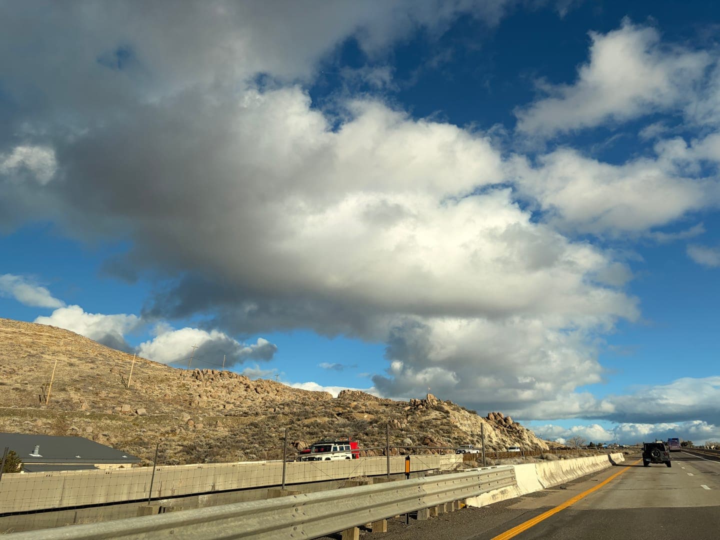 Driving along a highway with a clear view of a rugged, rocky hillside under a dynamic sky filled with both bright white and darker grey clouds. The road stretches ahead with a guardrail on the left, and a few vehicles are visible in the distance, including a black Jeep. The scene captures the expansive feeling of a road trip through a natural, somewhat arid landscape.