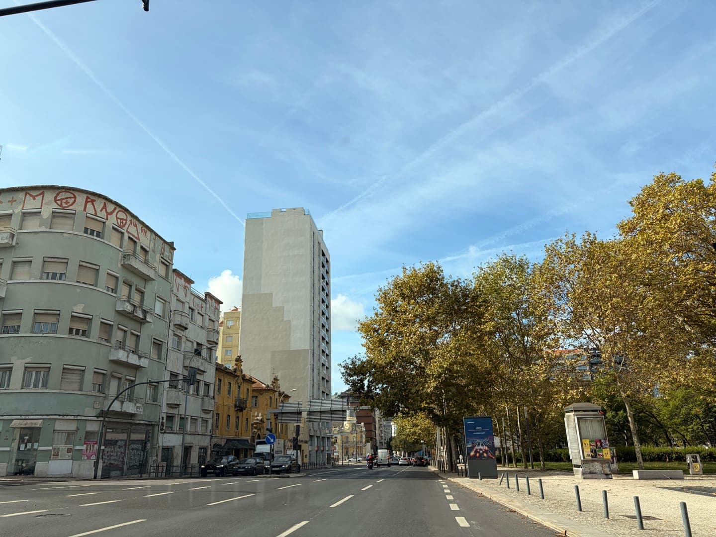 A wide-angle shot captures a bustling city street under a bright blue sky with wispy clouds and contrails. On the left, an older building with graffiti on its roof, including the word 'MORNO,' stands next to a modern high-rise. To the right, a line of trees with golden-brown leaves borders a park, adding a touch of nature to the urban landscape.