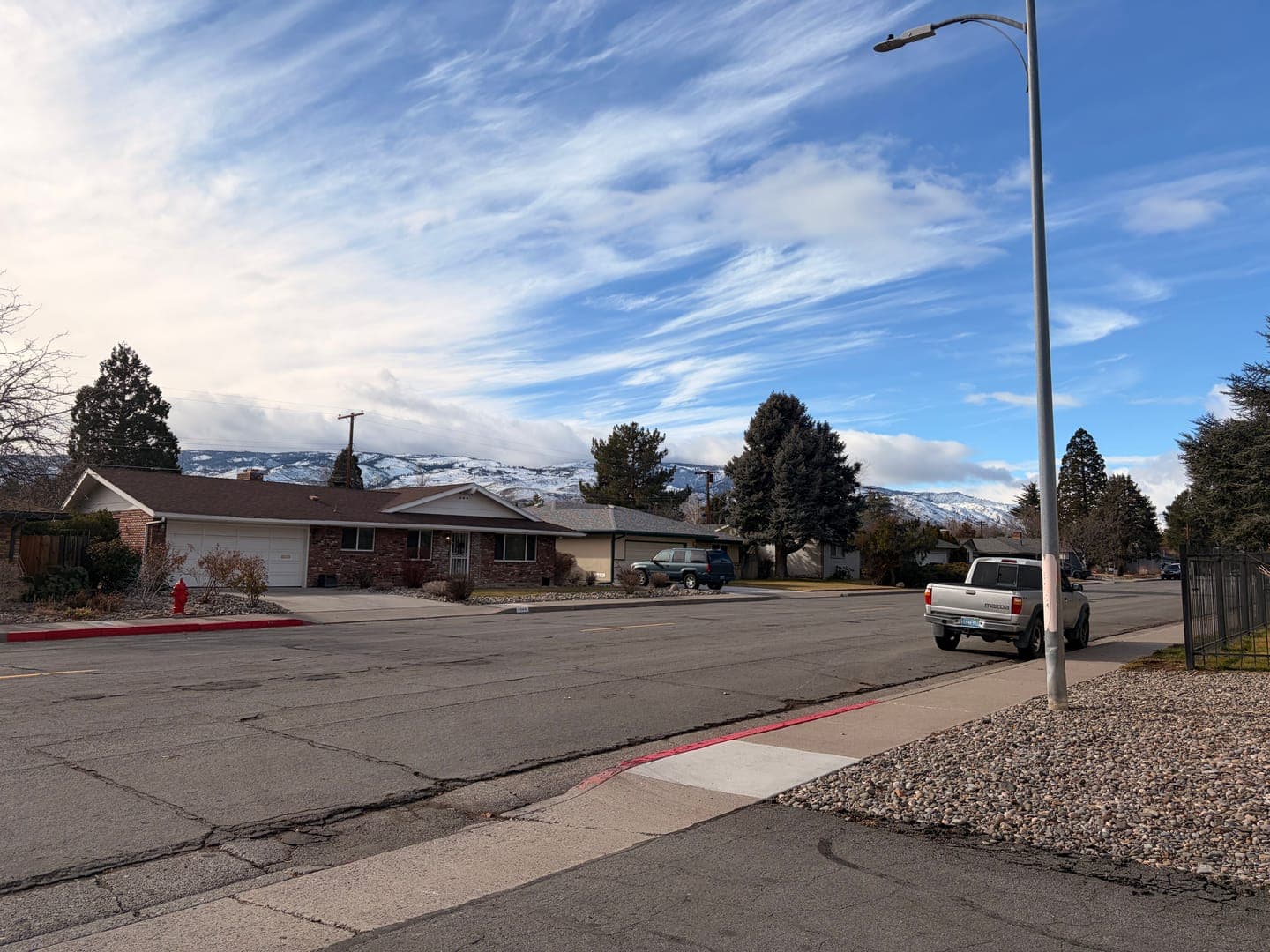 A quiet suburban street unfolds under a dramatic sky, with wispy clouds stretching across a vibrant blue. In the foreground, a brick house with a brown roof sits comfortably, while a silver pickup truck is parked on the side of the road, near a tall streetlamp. Snow-capped mountains form a majestic backdrop, adding a touch of natural grandeur to this peaceful neighborhood scene.