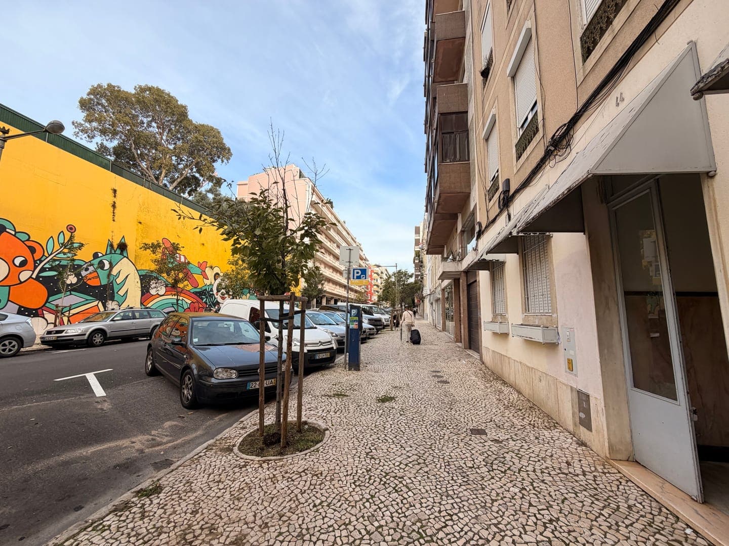A vibrant street scene unfolds with a striking yellow wall adorned with colorful graffiti on the left, contrasting with the more subdued tones of the buildings on the right. Parked cars line the street, and a person with a rolling suitcase walks along the patterned cobblestone sidewalk, adding a touch of everyday life to the urban landscape. The bright sky overhead suggests a pleasant day, making for an engaging visual blend of art, architecture, and daily activity.