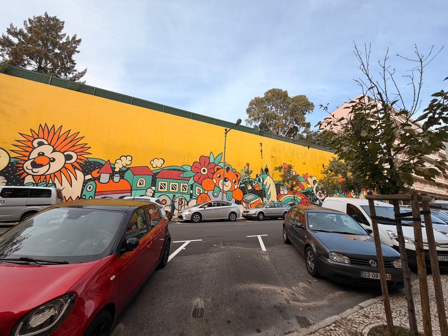 This vibrant street art transforms a plain wall into a whimsical wonderland, featuring a cheerful lion, a colorful train, and other playful characters. The bright yellow backdrop and cartoonish style create a joyful and imaginative atmosphere, bringing a splash of fun to the urban landscape. Parked cars line the street in front of this delightful mural, adding a touch of everyday life to the fantastical scene.