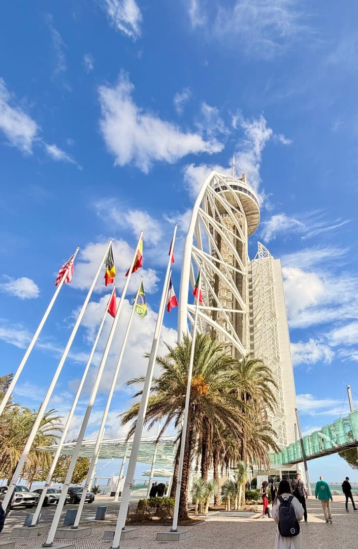 Soaring high above the vibrant city, the Vasco da Gama Tower in Lisbon, Portugal, stands as a magnificent landmark against a backdrop of a clear blue sky dotted with fluffy white clouds. International flags flutter proudly in the foreground, adding a touch of global charm to the scene, while lush palm trees frame the base of the tower, hinting at the city's warm climate. This architectural marvel, with its distinctive spiral design, invites visitors to explore its heights and enjoy breathtaking panoramic views of the Tagus River and beyond.