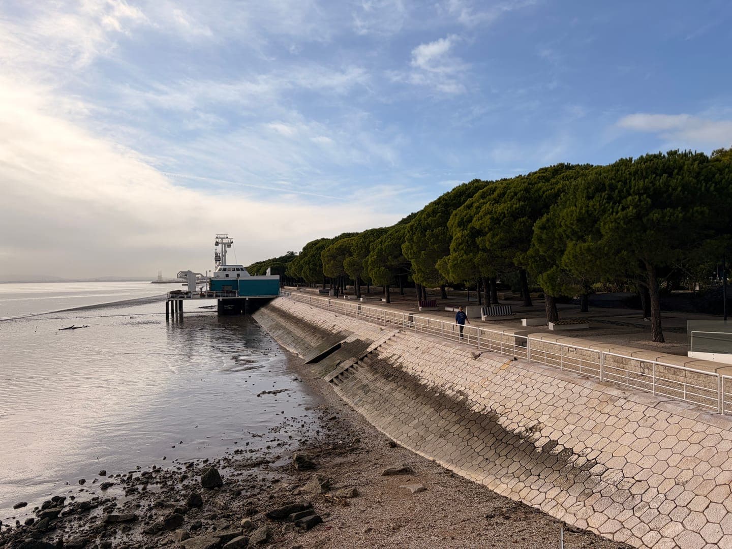 A serene waterfront scene unfolds under a partly cloudy sky, featuring a unique teal and white building on stilts extending over the water. A paved promenade lined with lush green trees stretches along the shore, where a lone figure enjoys a peaceful stroll. The calm waters reflect the soft light, creating a tranquil atmosphere perfect for a leisurely day by the river.
