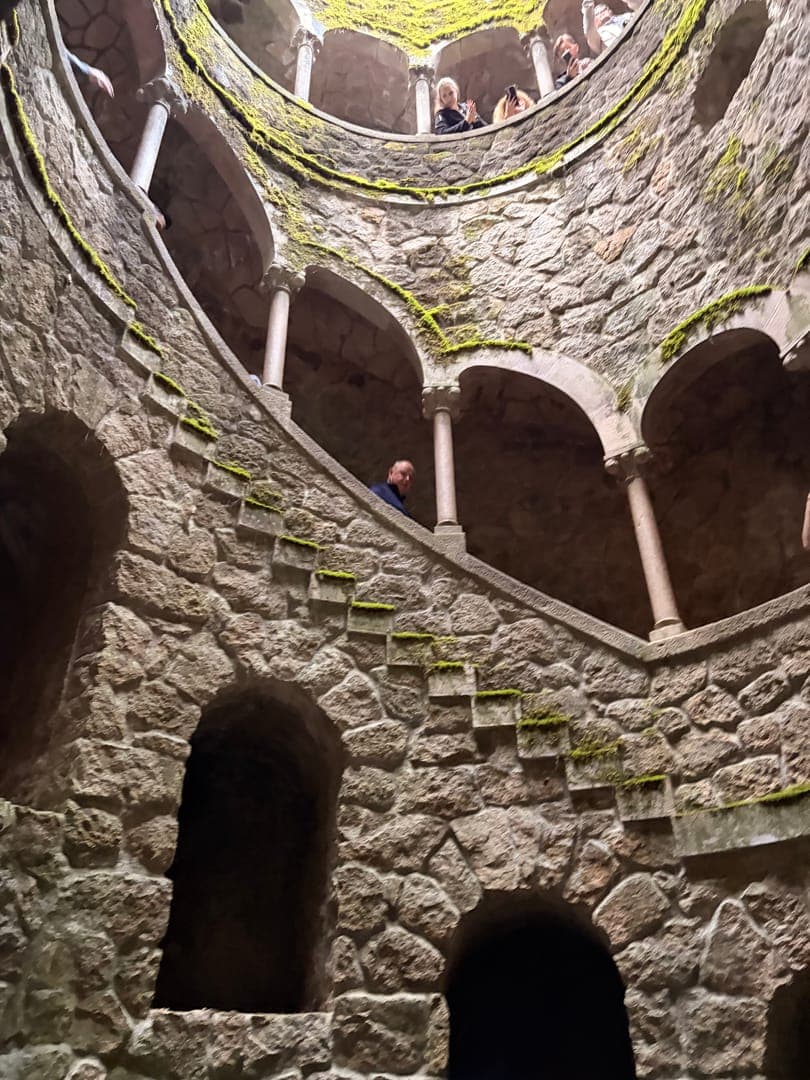 Step into the enchanting depths of the Initiation Well at Quinta da Regaleira, where a spiral staircase descends into mystery. The moss-covered stone walls and intricate arches create an otherworldly atmosphere, inviting exploration. Glimpses of visitors on the upper levels add a sense of scale to this captivating architectural wonder.