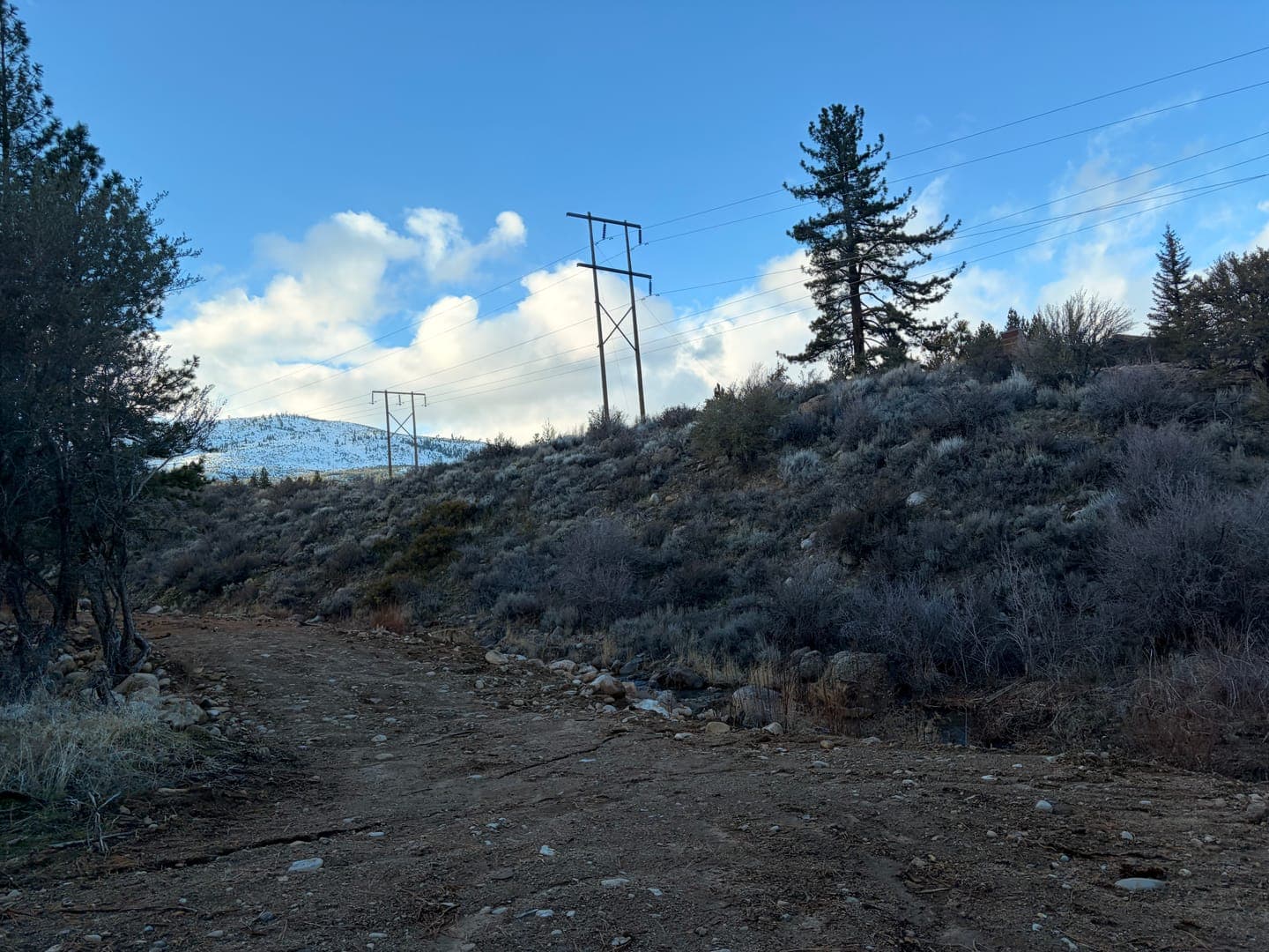 A dirt road winds through a rugged, hilly landscape under a bright blue sky dotted with fluffy white clouds. Power lines stretch across the scene, leading towards a snow-capped mountain in the distance, adding a touch of winter's embrace to the otherwise earthy tones of the foreground. The sparse vegetation and clear sky evoke a sense of quiet solitude and natural beauty.