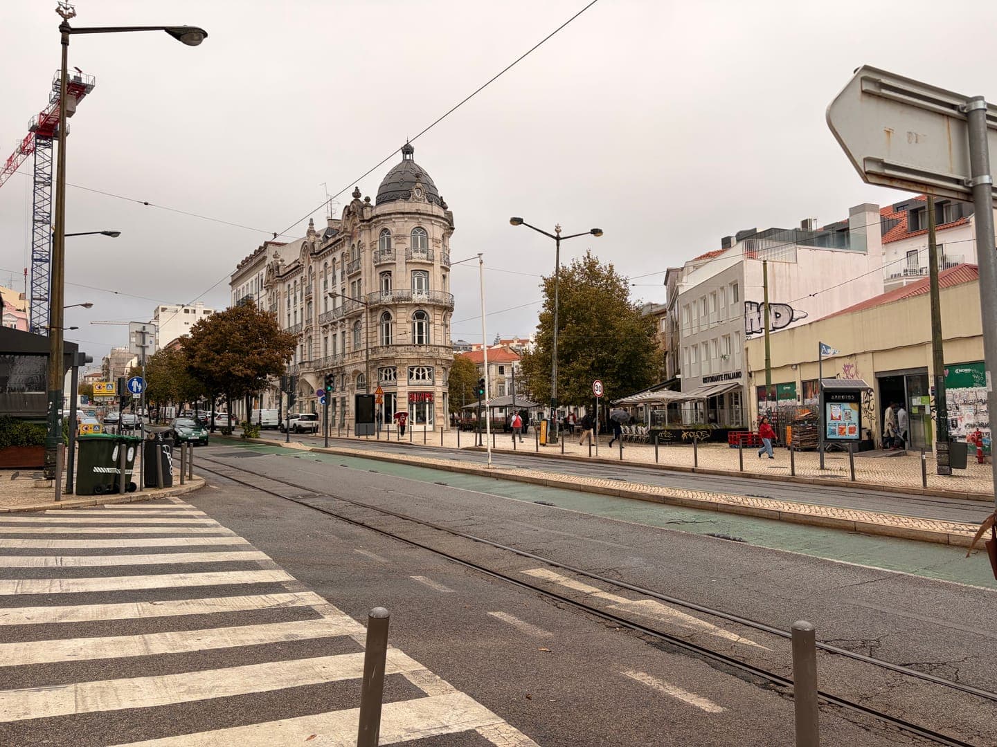 A wide-angle shot captures a bustling city street on an overcast day, with a prominent, ornate building featuring a domed roof standing out on the left. Tram tracks run down the center of the road, flanked by sidewalks where people stroll past shops and cafes. The scene is a vibrant blend of historic architecture and modern urban life, hinting at the everyday rhythm of a European city.