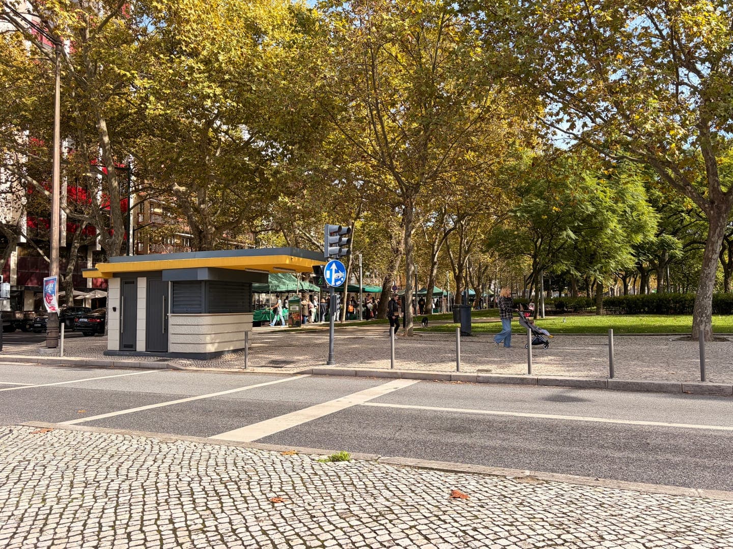 A charming kiosk with a yellow roof stands beside a bustling street, its modern design contrasting with the traditional cobblestone pavement. Lush green and golden-leaved trees line the park across the road, where people stroll and a market with green awnings hints at local delights. The scene captures a vibrant urban park on a sunny day, blending city life with natural beauty.