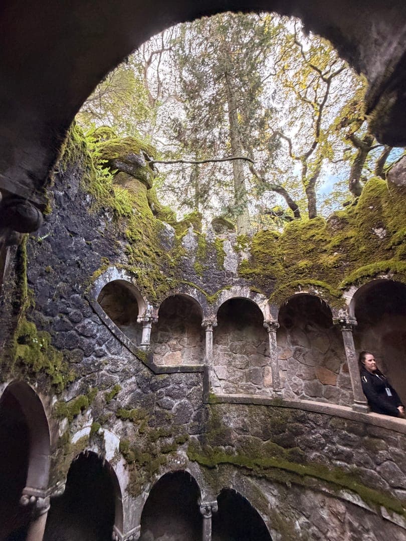 Step into a fairytale at Quinta da Regaleira's Initiation Well, where ancient stone walls draped in vibrant moss lead to a glimpse of the sky above. This enchanting spiral staircase, adorned with intricate arches, evokes a sense of mystery and wonder. A lone figure rests on a ledge, adding a human element to this magical, otherworldly scene.