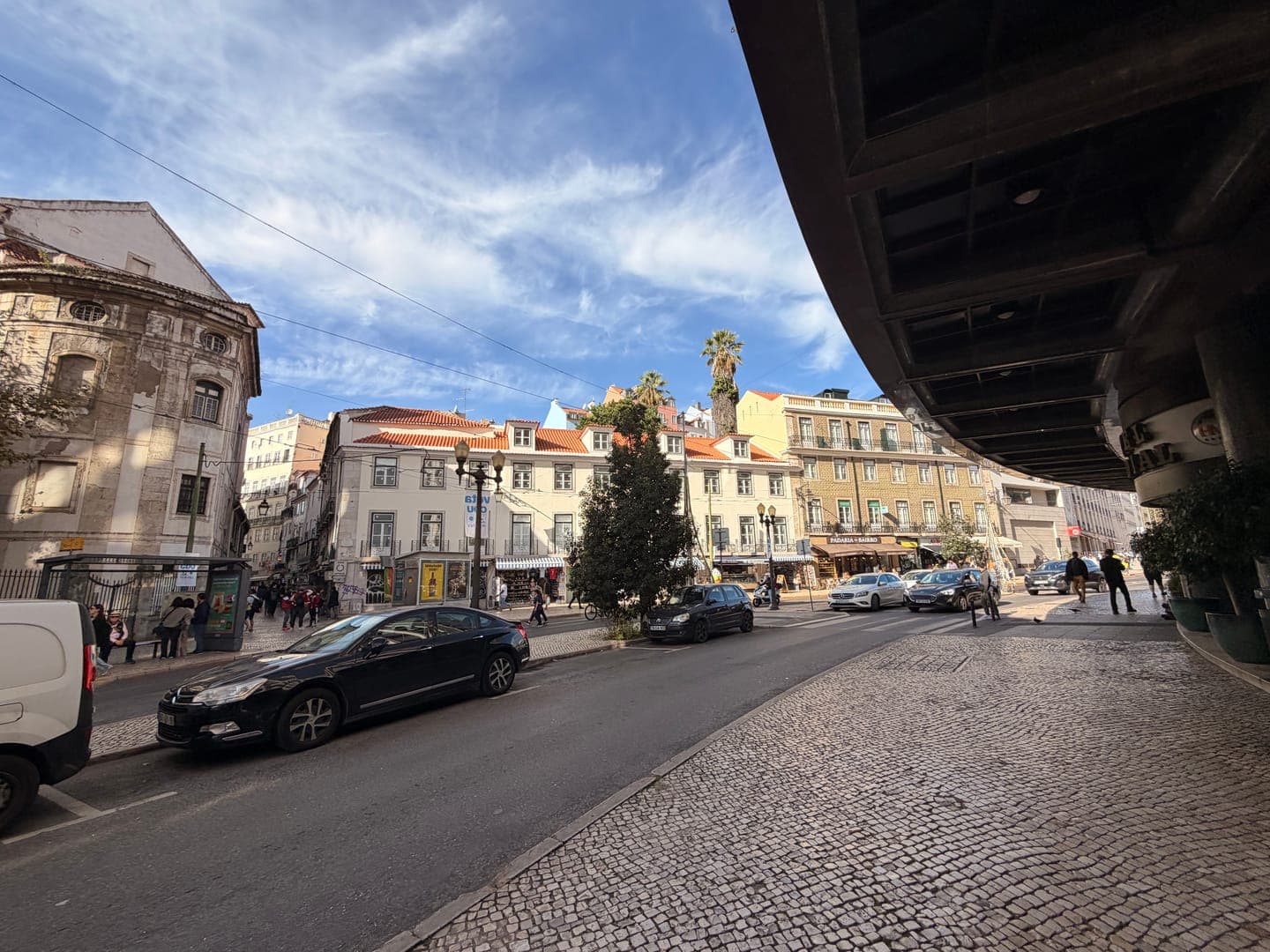This vibrant street scene captures the essence of a bustling European city, likely Lisbon, with its charming architecture and lively atmosphere. The cobblestone street in the foreground leads the eye towards a row of colorful buildings, some with traditional red-tiled roofs, under a bright blue sky dotted with wispy clouds. Cars line the street and people stroll along the sidewalks, adding to the dynamic feel of this picturesque urban landscape.