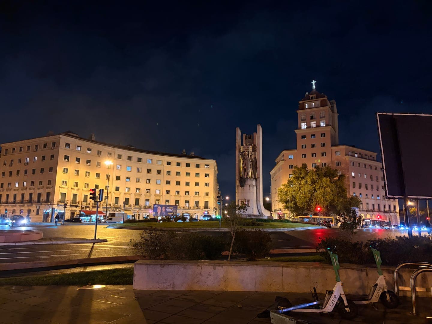 Night falls over a bustling city square, where the warm glow of streetlights and building windows illuminates the scene. A prominent monument stands tall in the center, flanked by a curved building on the left and a taller structure with a cross atop its roof on the right. The dark sky, hints of traffic lights, and parked Lime scooters add to the urban ambiance of this evening shot.