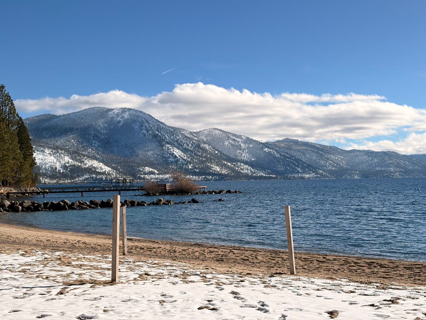 Winter's embrace at Lake Tahoe paints a stunning scene, with snow-dusted mountains rising majestically against a bright blue sky. The calm, deep blue waters of the lake reflect the serene beauty, while a sandy beach in the foreground shows remnants of snow, hinting at the season's chill. A distant pier and rocky shoreline add texture to this picturesque landscape, inviting contemplation and appreciation for nature's artistry.