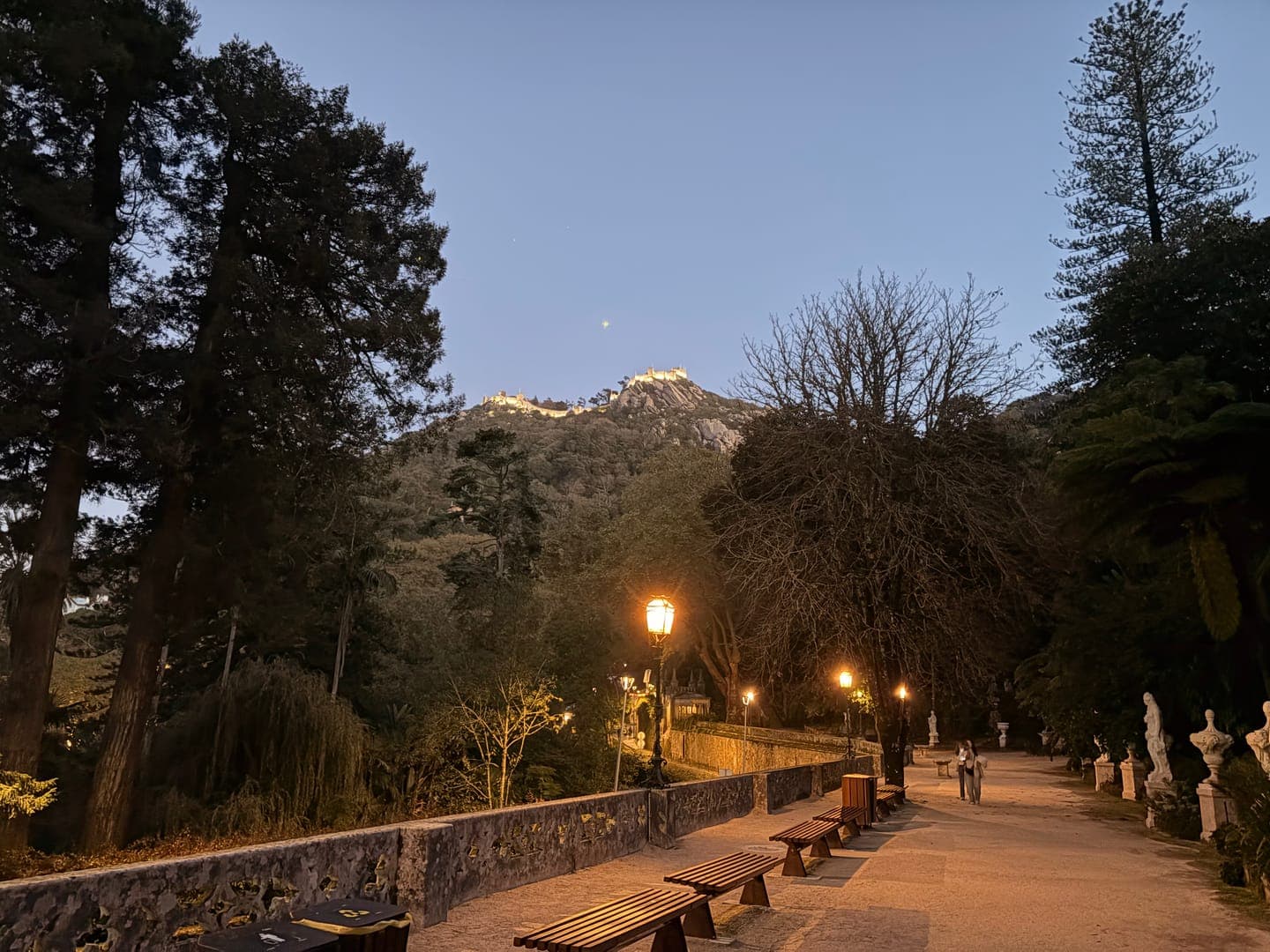 As dusk settles, a serene pathway illuminated by warm streetlights invites a leisurely stroll through a lush park. In the distance, a majestic castle perched atop a mountain glows, adding a touch of enchantment to the twilight scene. The tranquil atmosphere, with its blend of natural beauty and historical grandeur, creates a perfect setting for an evening escape.