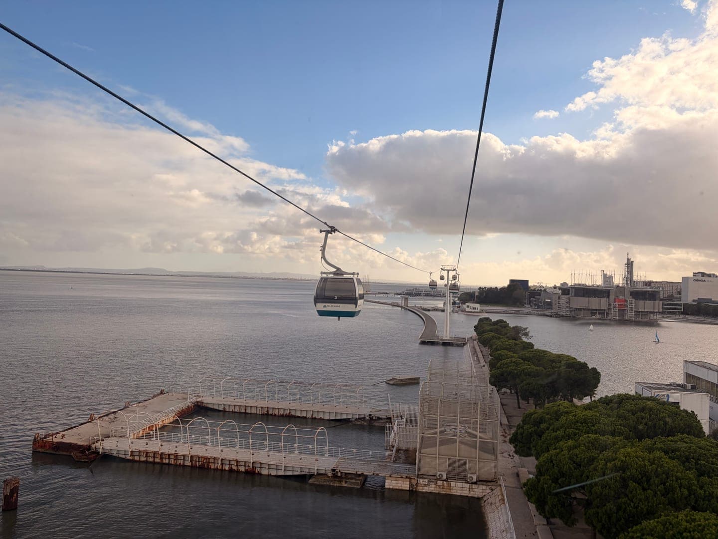 A cable car glides over the water, offering a scenic view of a city's waterfront. The sky is a mix of blue and cloudy, casting a soft light over the urban landscape and the calm body of water. In the distance, buildings line the shore, and a long pier stretches out, adding to the tranquil yet developed atmosphere.