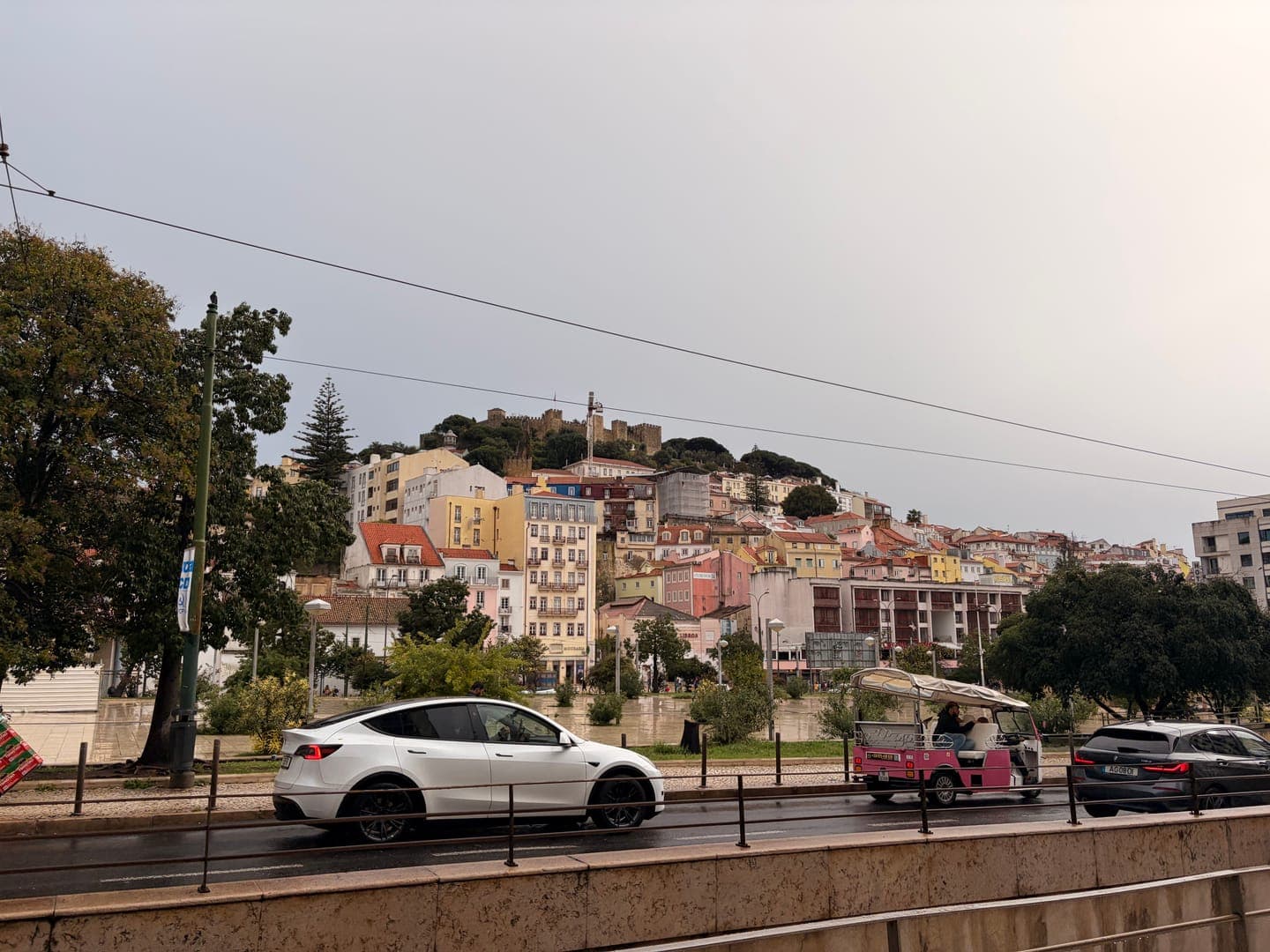 A vibrant cityscape unfolds under a soft, overcast sky, with colorful buildings cascading down a hill topped by a historic castle. In the foreground, a modern white car and a charming pink tuk-tuk share the road, hinting at a blend of old and new transportation in this bustling urban setting. The scene captures a lively street view, offering a glimpse into the daily rhythm of a picturesque European city.