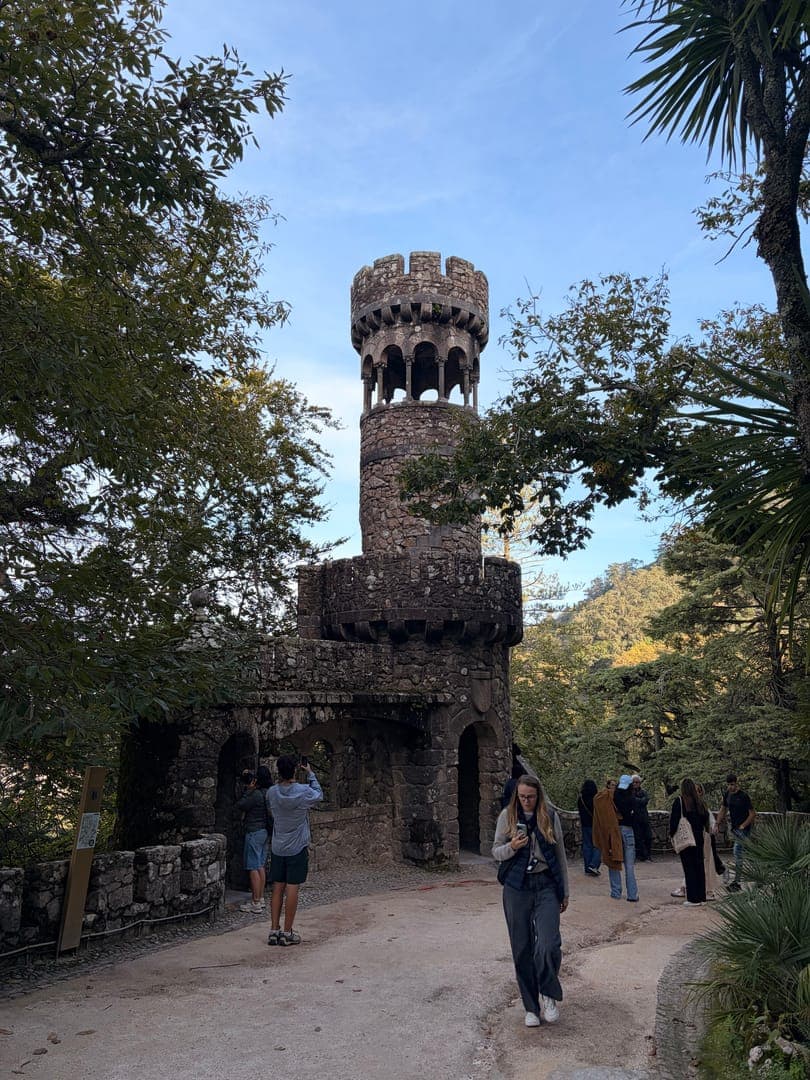 Exploring the enchanting Quinta da Regaleira, this image captures the iconic Initiation Well tower peeking through lush greenery. The ancient stone structure, with its intricate details, evokes a sense of mystery and historical grandeur. Visitors wander along the path, soaking in the magical atmosphere of this Portuguese wonder.