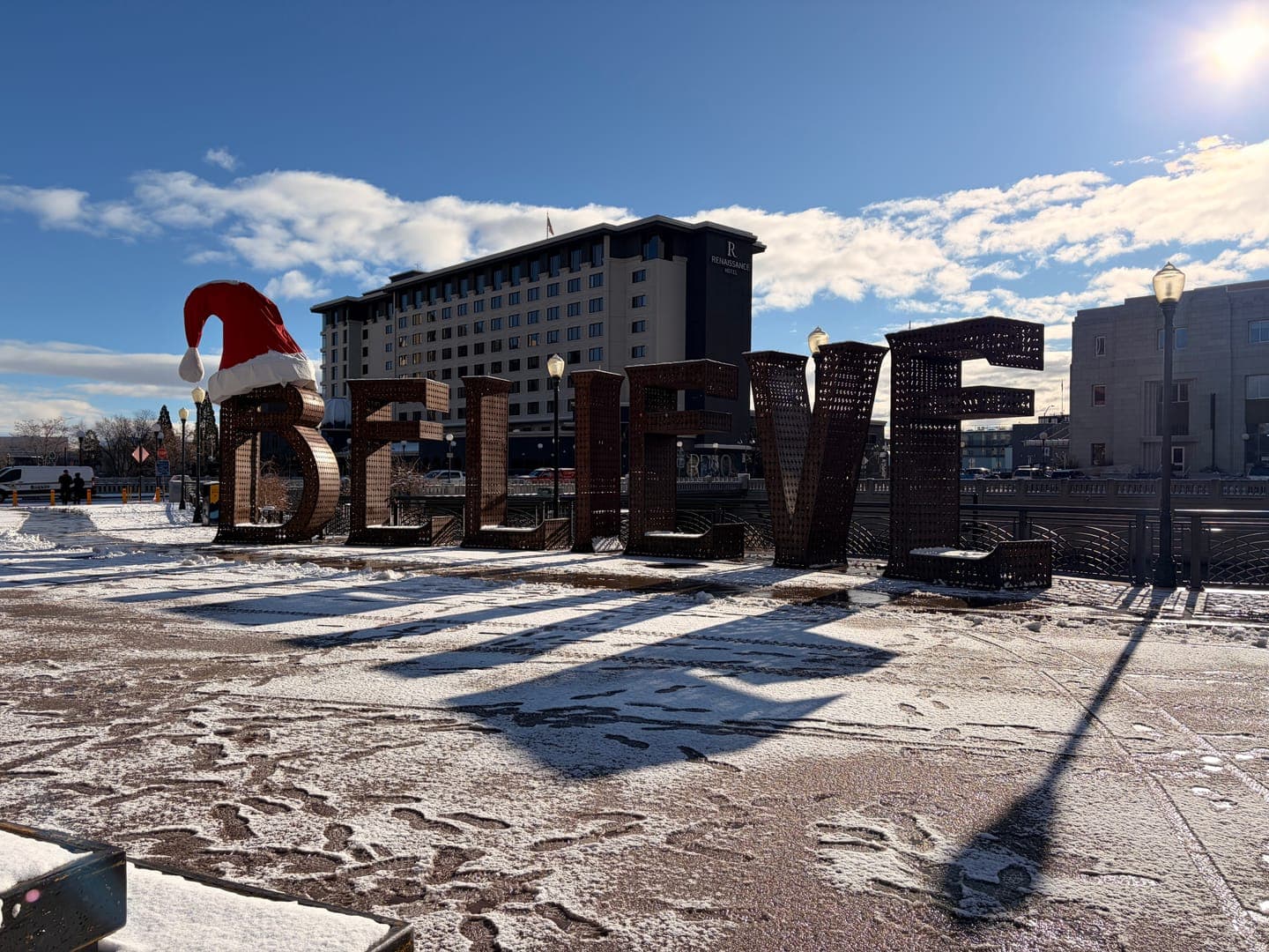 Embrace the festive spirit with this charming outdoor display! Large, rustic letters spell out 'BELIEVE,' with a cheerful Santa hat perched atop the 'B,' all dusted with a light layer of snow. The bright winter sun casts long shadows, adding a touch of magic to this holiday scene.