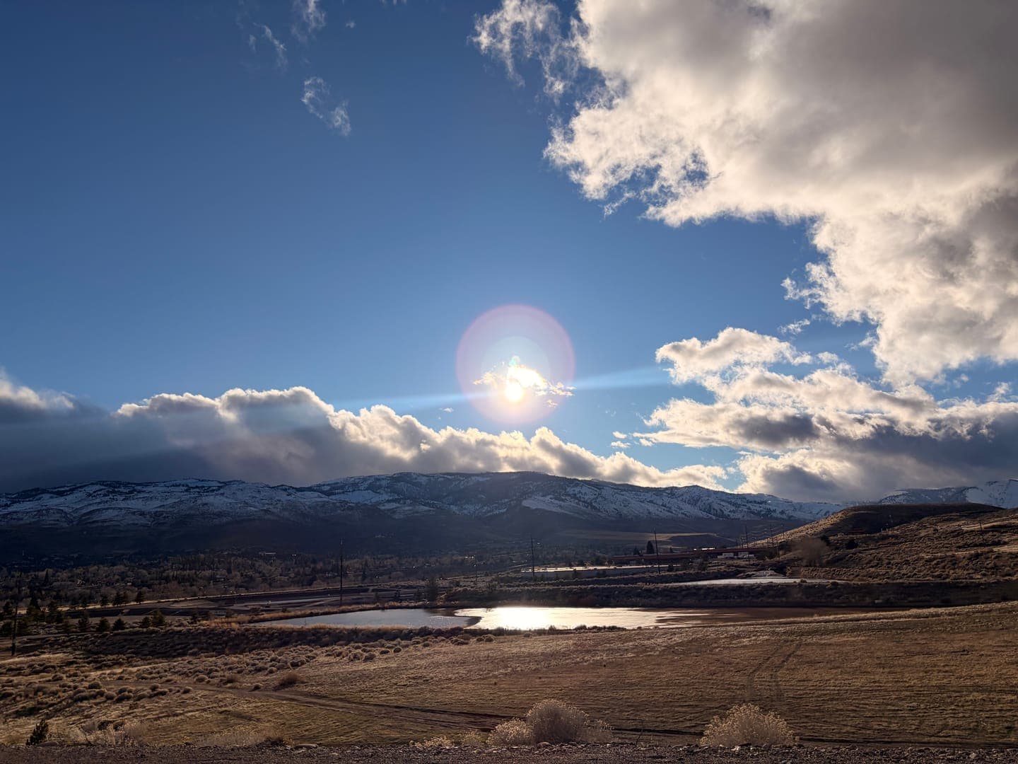 A vibrant sunburst pierces through a partly cloudy blue sky, casting a warm glow over a serene landscape. Snow-capped mountains rise in the distance, overlooking a tranquil body of water reflecting the bright sunlight, surrounded by dry, golden fields. The scene evokes a peaceful, expansive feeling of nature's beauty under a dynamic sky.