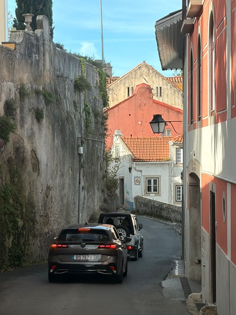 Winding through a charming, narrow street, two cars navigate the historic architecture of what appears to be a European town. The warm hues of the buildings, with their terracotta roofs and weathered walls, create a picturesque and inviting atmosphere. The scene captures a blend of old-world charm and modern life, with the cars adding a contemporary touch to the timeless setting.