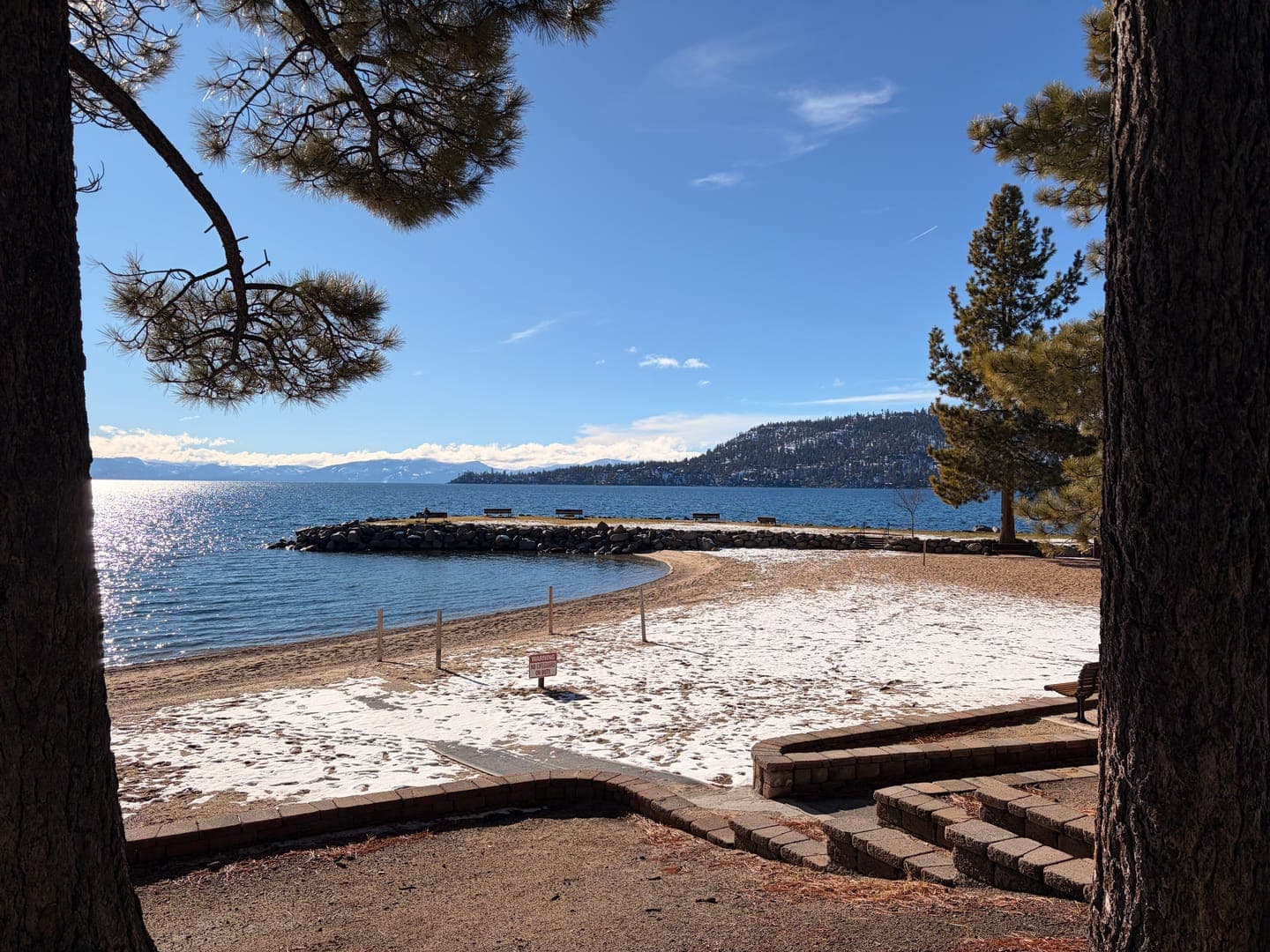 A serene winter scene unfolds at the edge of a lake, where patches of snow dust the sandy beach. The calm blue water reflects the bright sky, with distant snow-capped mountains adding to the picturesque backdrop. Framed by the dark trunks and branches of pine trees, the image captures a peaceful moment where winter meets the water's edge.