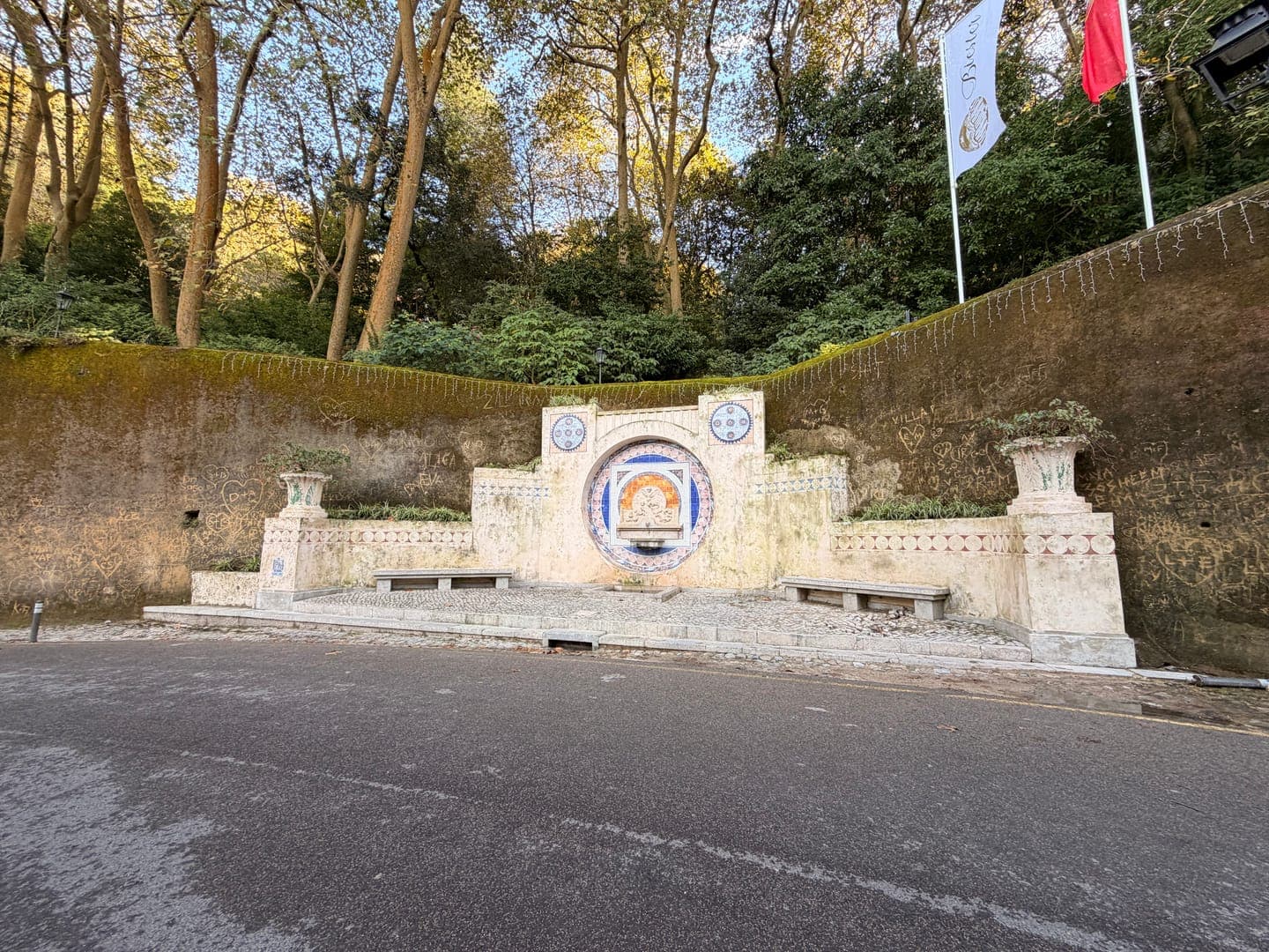 This charming image captures a beautifully tiled fountain built into a moss-covered wall, nestled at the base of a lush, tree-filled hillside. Two stone benches flank the central fountain, inviting visitors to rest and enjoy the serene atmosphere. Above, a white flag with 'Beater' visible and a red flag flutter against the clear sky, adding a touch of color to this picturesque scene.