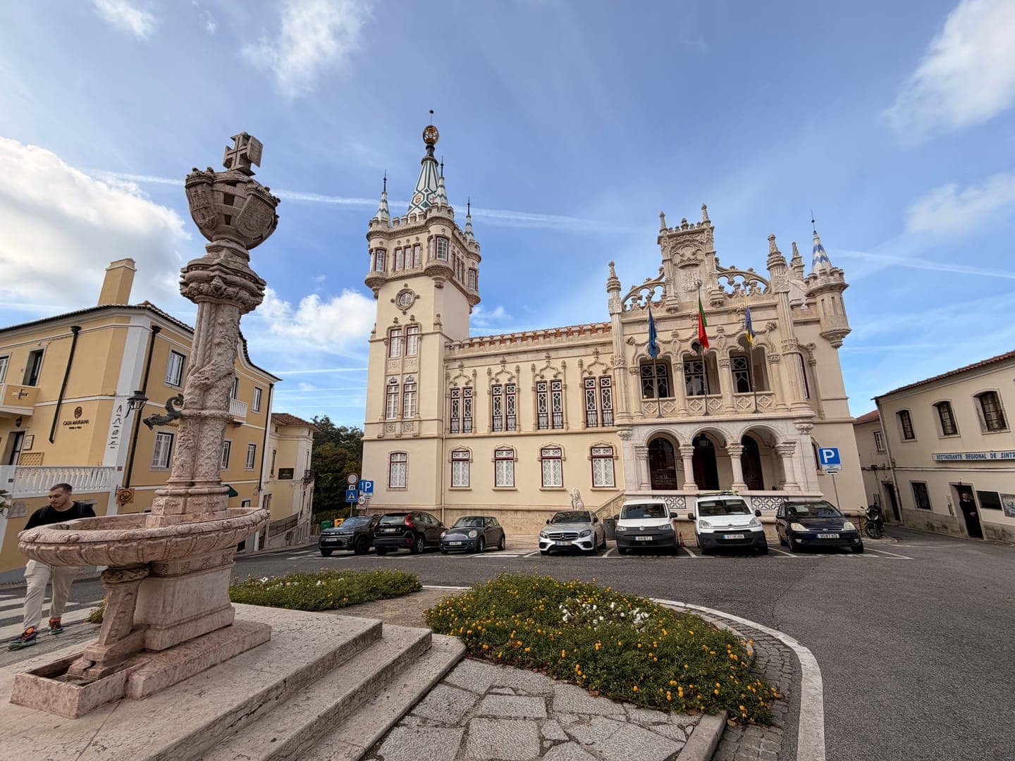 A grand, ornate building with flags flying proudly stands under a bright sky, its intricate architecture a focal point of the town square. In the foreground, a historic stone fountain adds to the charm, while parked cars and a pedestrian hint at the daily life unfolding around this impressive structure. The scene captures a blend of historical grandeur and everyday activity, inviting viewers to imagine strolling through this picturesque European town.