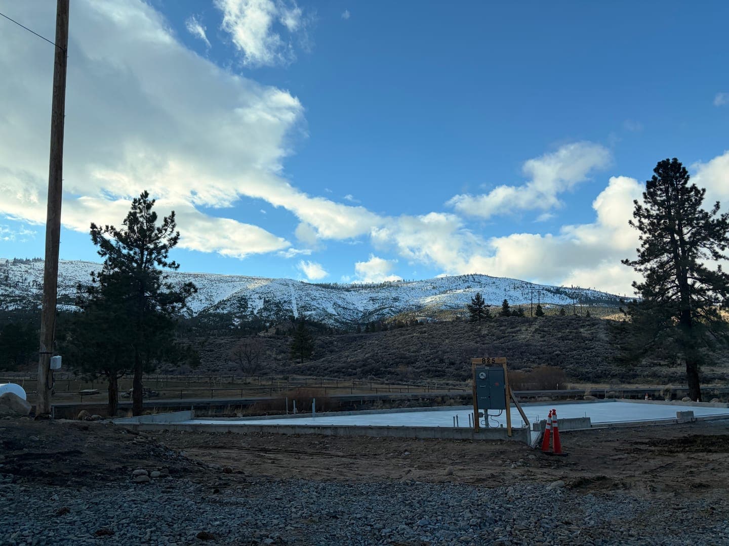 A new beginning unfolds against a stunning winter backdrop! This image captures a freshly poured concrete foundation, ready for construction, with a majestic snow-capped mountain range in the distance under a bright blue sky dotted with fluffy clouds. The scene is framed by tall pine trees and a utility pole, hinting at a blend of nature and development in this serene landscape.
