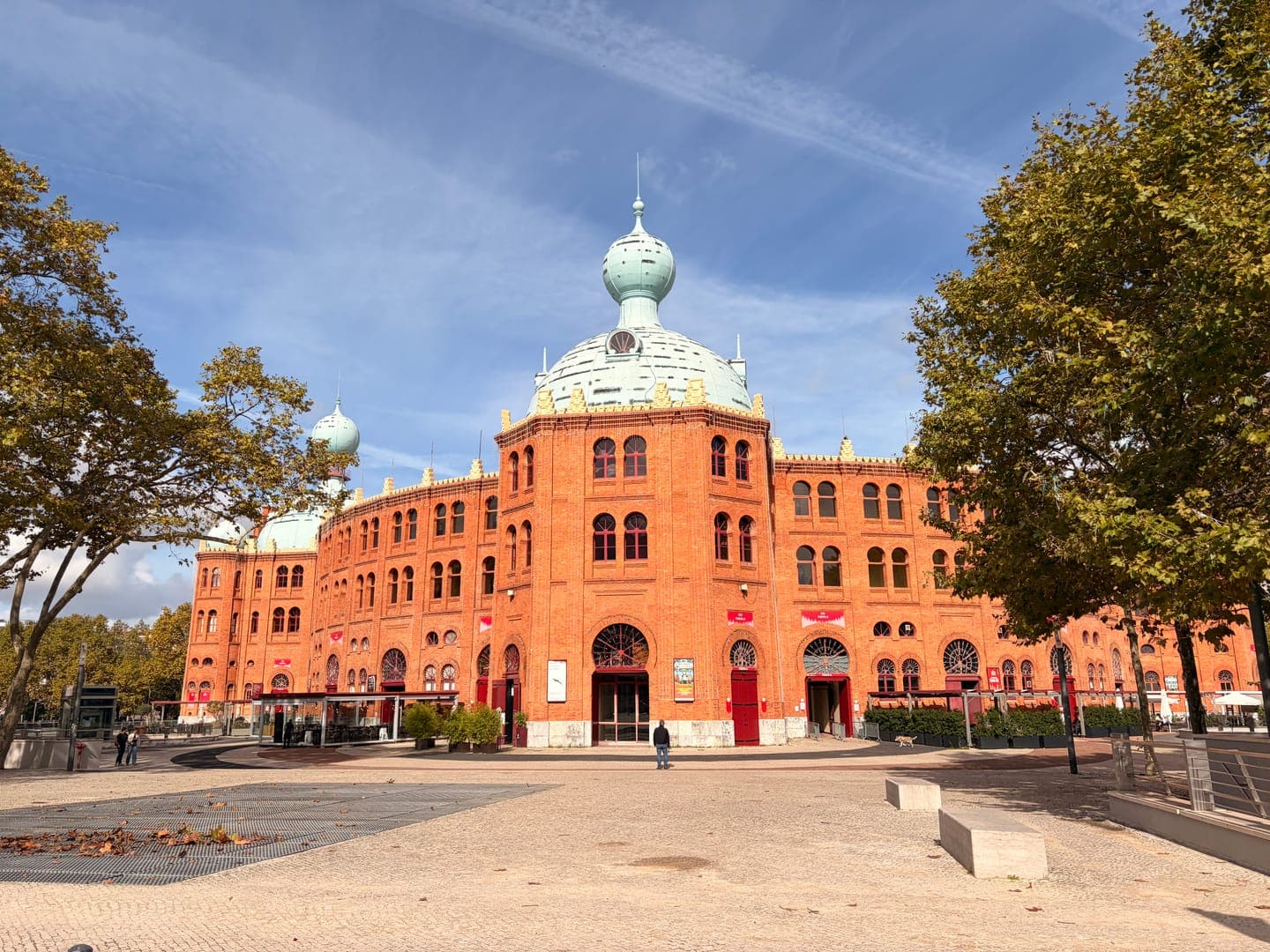 The Campo Pequeno bullring in Lisbon, Portugal, stands majestically under a bright blue sky, its distinctive red brick and light blue domes creating a striking contrast. Lush green trees frame the historic building, adding a touch of nature to the urban landscape. The grand architecture and clear weather evoke a sense of timeless beauty and cultural richness.