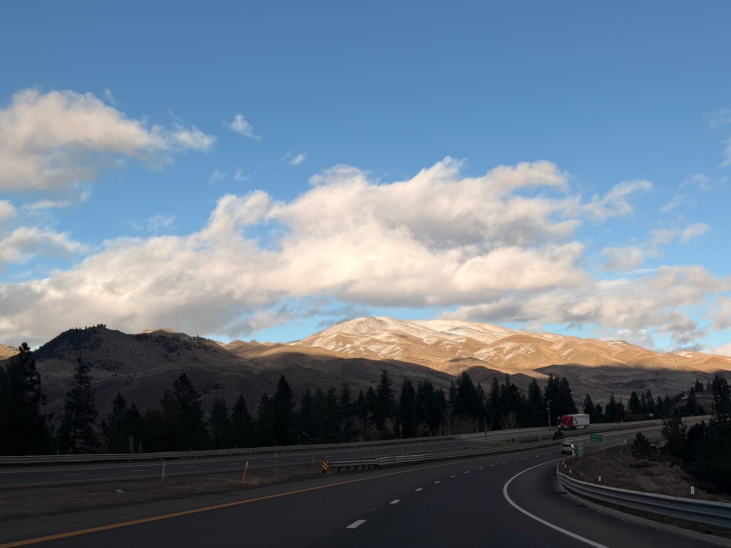 Driving through a scenic landscape, a highway curves alongside a range of mountains, some of which are dusted with snow. The sky above is a vibrant blue, dotted with fluffy white clouds, and the warm sunlight illuminates parts of the mountains, creating a beautiful contrast with the shadowed areas. Pine trees line the base of the mountains, adding to the serene and picturesque view.