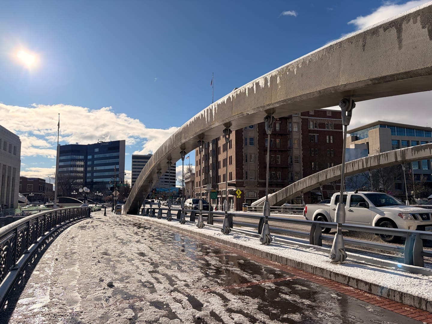 A sunny winter day illuminates a city bridge, its concrete arches adorned with icicles, hinting at recent cold weather. The pedestrian walkway and road below are covered in a mix of melting snow and ice, reflecting the bright sky. In the background, modern and older buildings stand under a partly cloudy sky, with a white pickup truck visible on the bridge.