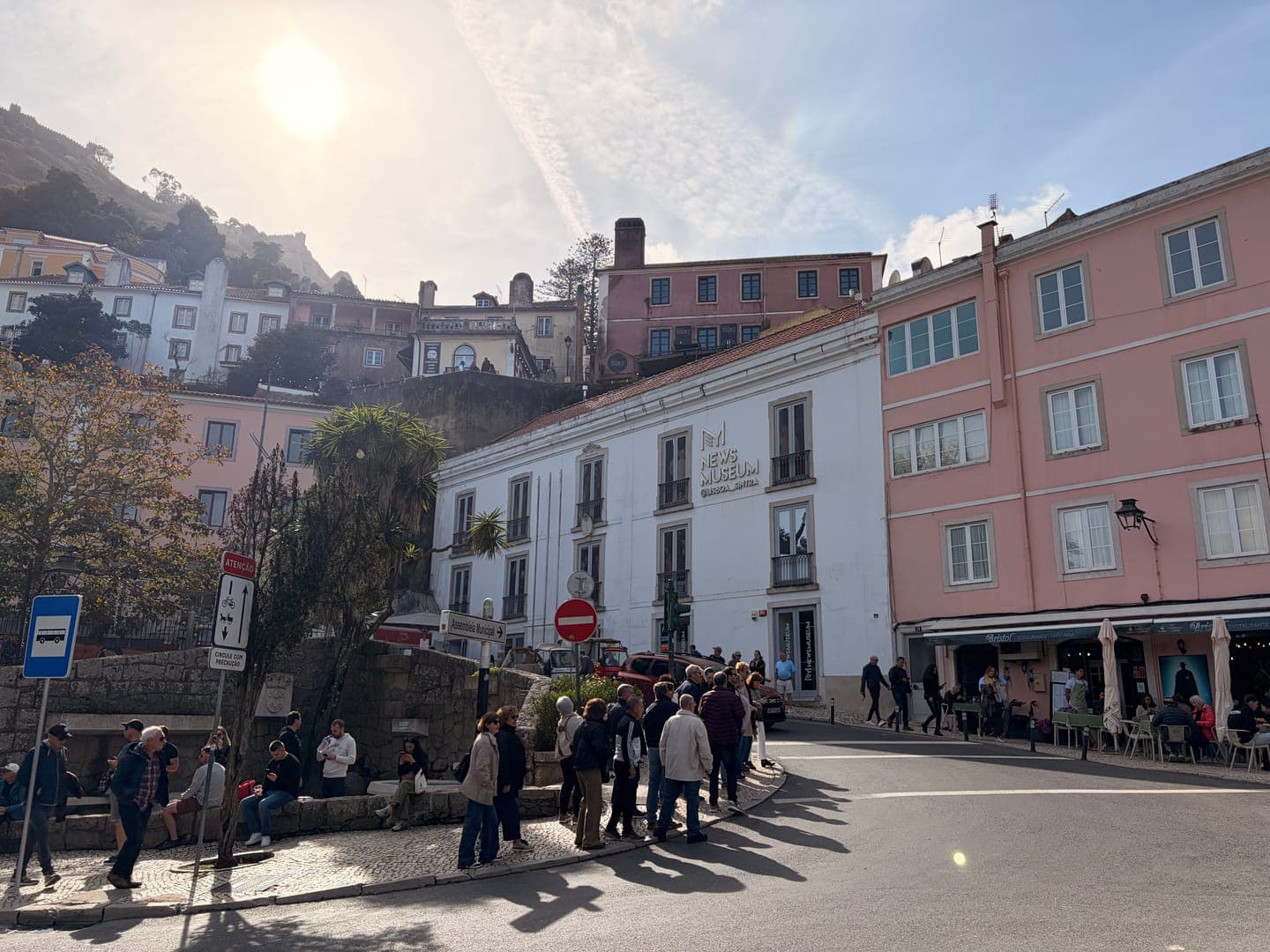 A bustling street scene unfolds in a charming European town, with people gathered on the sidewalk and a bus stop sign visible. The architecture is a delightful mix of styles, featuring a prominent white building labeled 'NEWS MUSEUM' and a soft pink building with outdoor seating. The bright sun casts long shadows, adding to the lively atmosphere of this picturesque location.