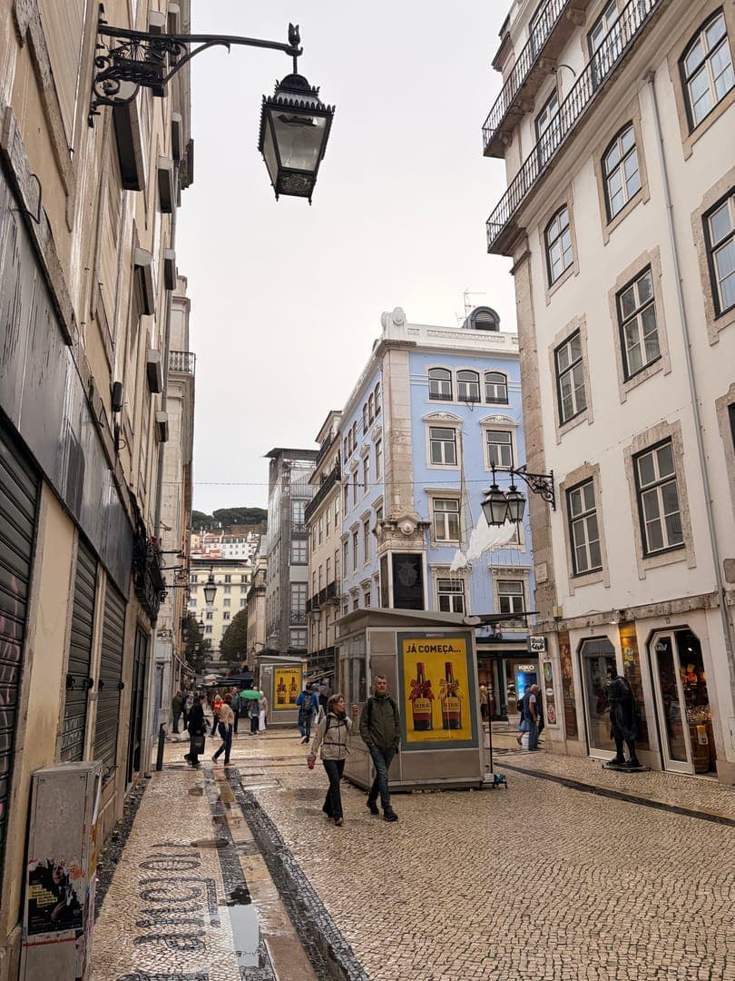 Strolling through a charming European street, the cobblestone path, still damp from a recent rain, reflects the soft light of the overcast sky. Ornate street lamps extend from the classic architecture, with one prominent lamp hanging overhead, adding to the old-world ambiance. In the distance, a blue building stands out, while closer by, a kiosk with the text 'JÁ COMEÇA...' (It's starting...) hints at local promotions or events, as people leisurely walk along the historic lane.