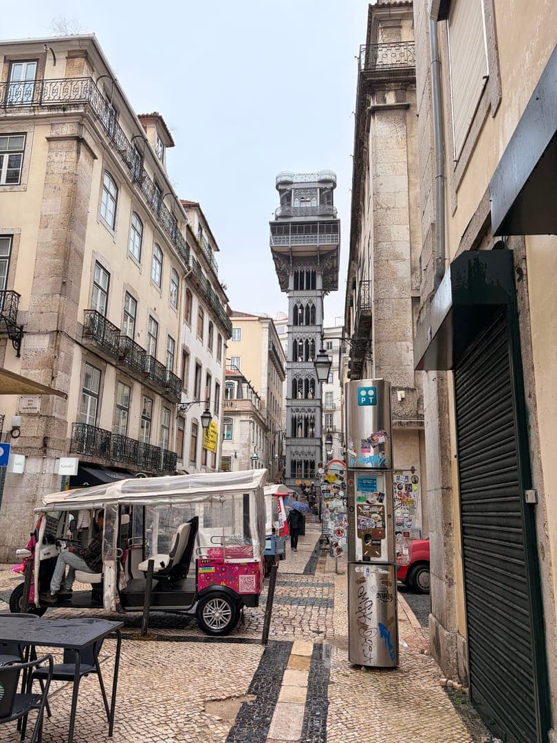 A charming street scene in Lisbon, Portugal, captures the iconic Santa Justa Lift towering in the background. The cobblestone street is lined with historic buildings and a colorful tuk-tuk awaits passengers, adding a vibrant touch to the old-world charm. The mood is a blend of historical grandeur and everyday urban life, inviting exploration of this beautiful city.
