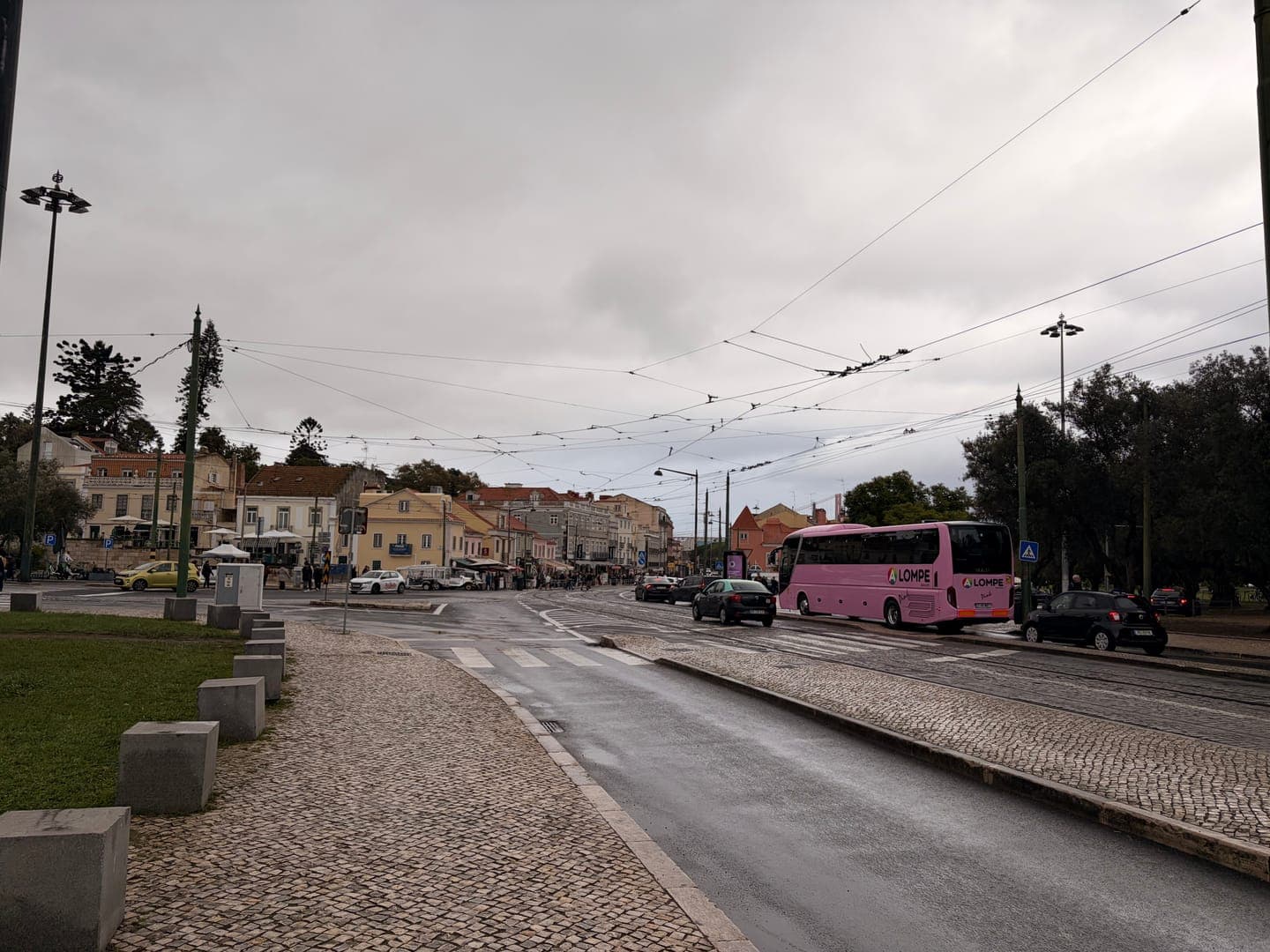 A vibrant pink 'LOMPE' bus stands out against the muted tones of a cloudy day in a bustling city street. The scene captures a typical urban landscape with historic buildings lining the background, cars navigating the wet roads, and a network of overhead wires crisscrossing the sky. The cobblestone sidewalk and grassy median add texture to this lively street view, hinting at a blend of old-world charm and modern transit.