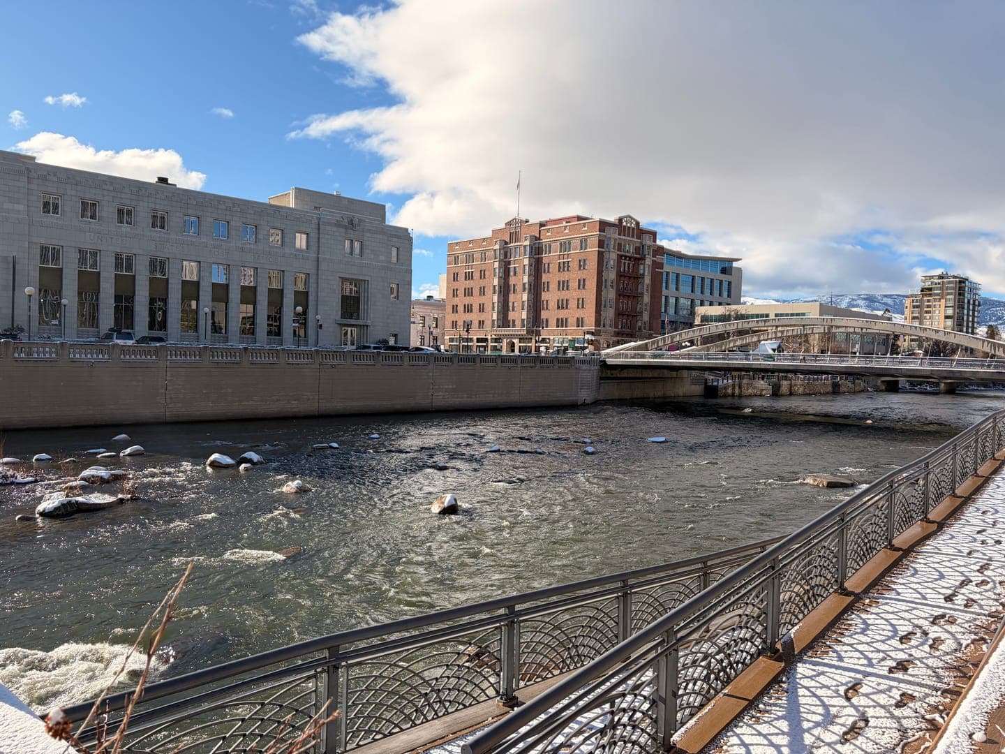 A scenic view of a river flowing through a city on a bright, cloudy day. The river, with snow-dusted rocks, is flanked by a modern building on the left and a historic brick building and a bridge on the right. A snowy walkway with decorative railings and footprints adds to the winter charm of the urban landscape.