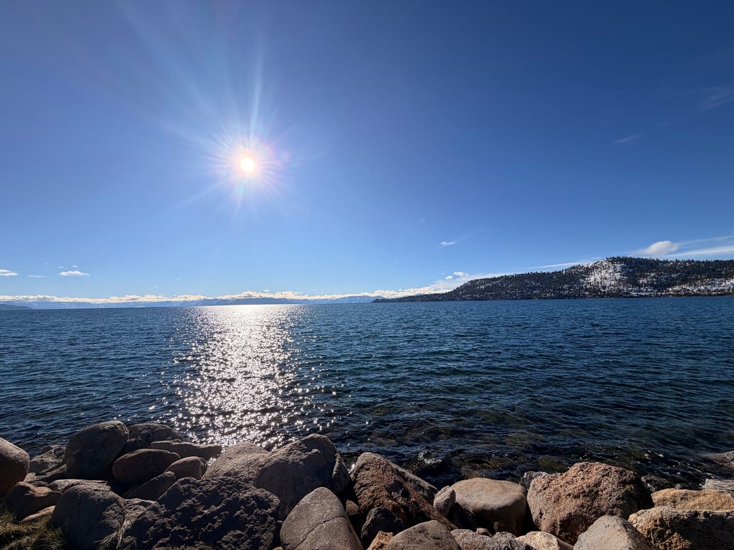 A bright sun shines over a vast body of water, creating a sparkling path of light across its surface. In the foreground, large rocks line the shore, while a distant, snow-dusted mountain range adds a majestic backdrop to the serene scene. The clear blue sky and calm waters evoke a peaceful and refreshing natural landscape.