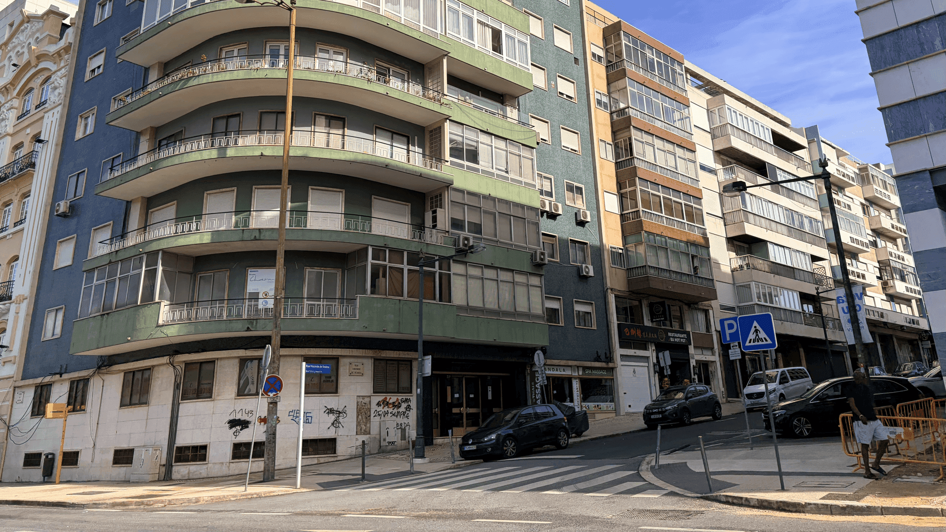 A vibrant street scene unfolds in Lisbon, Portugal, showcasing a mix of architectural styles under a bright sky. The prominent blue and green building with curved balconies adds a unique character to the urban landscape, while cars are parked along the street and a pedestrian crosses at a marked crosswalk. The image captures the lively atmosphere of a sunny day in the city.