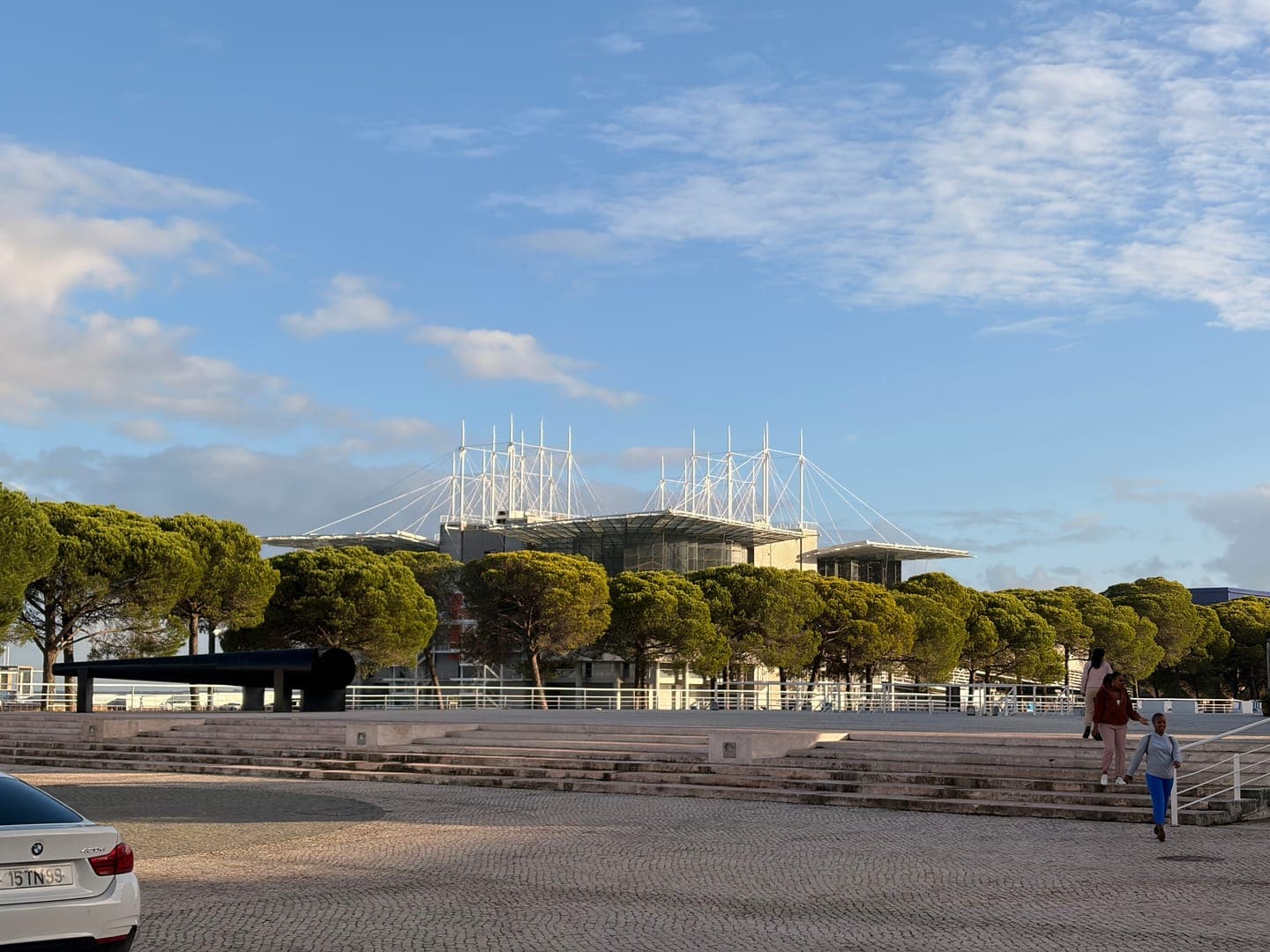 A modern building with a distinctive white, spiky roofline stands out against a bright blue sky with scattered clouds. Lush green trees line the foreground, partially obscuring the building, while a paved area with steps and a white car in the bottom left corner adds to the urban landscape. The scene evokes a sense of contemporary architecture nestled within a green, open space.